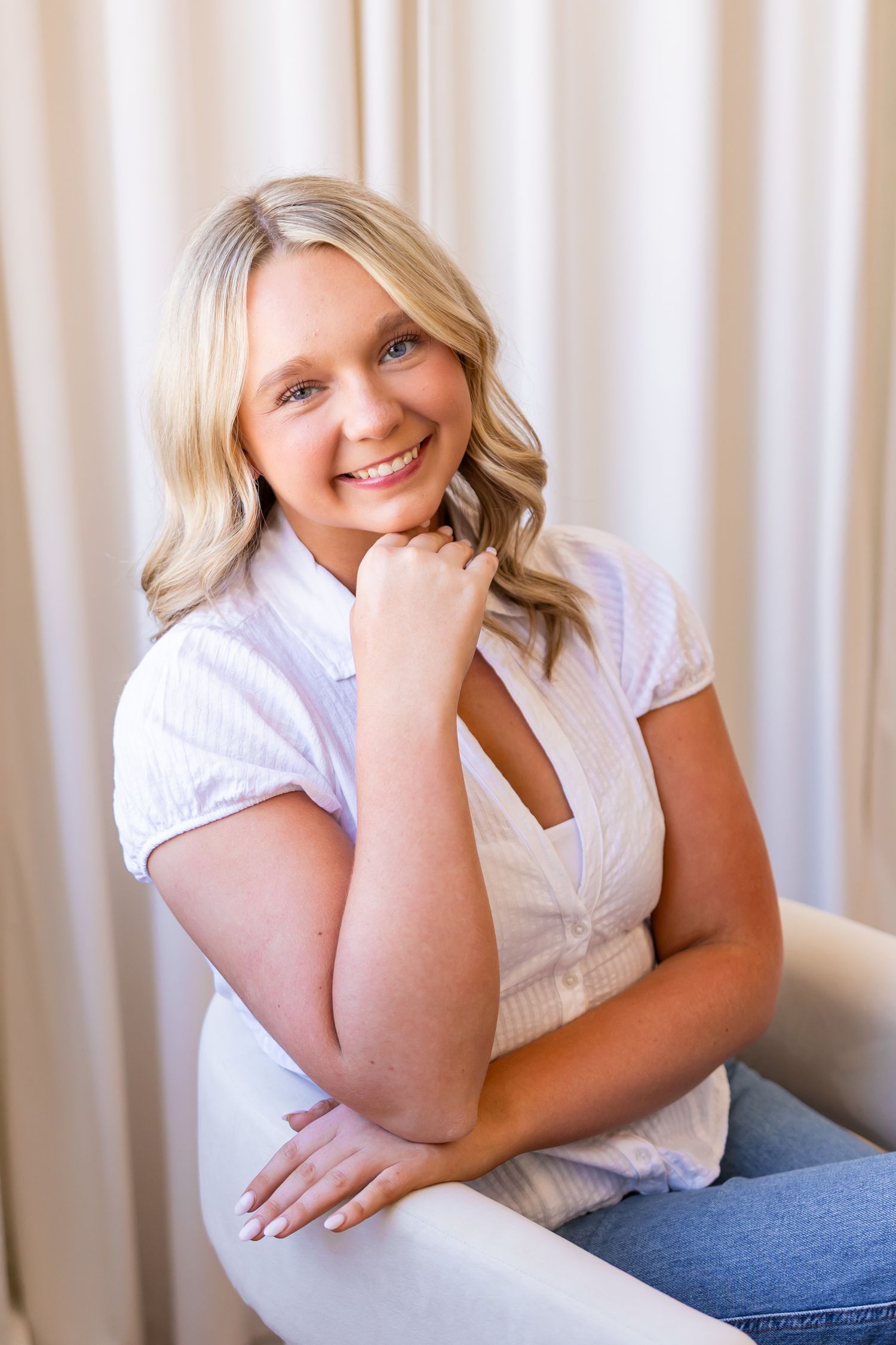 A smiling young woman with long blonde curly hair sits on a chair in a light-colored room.