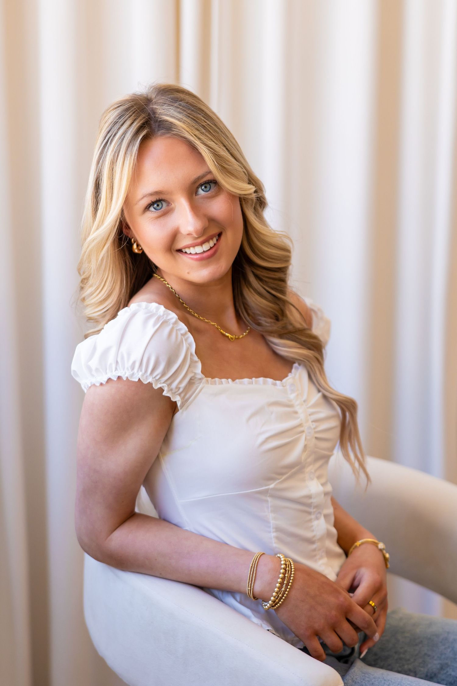 A smiling young woman with long blonde hair wearing a white top sits on a chair against a white background.