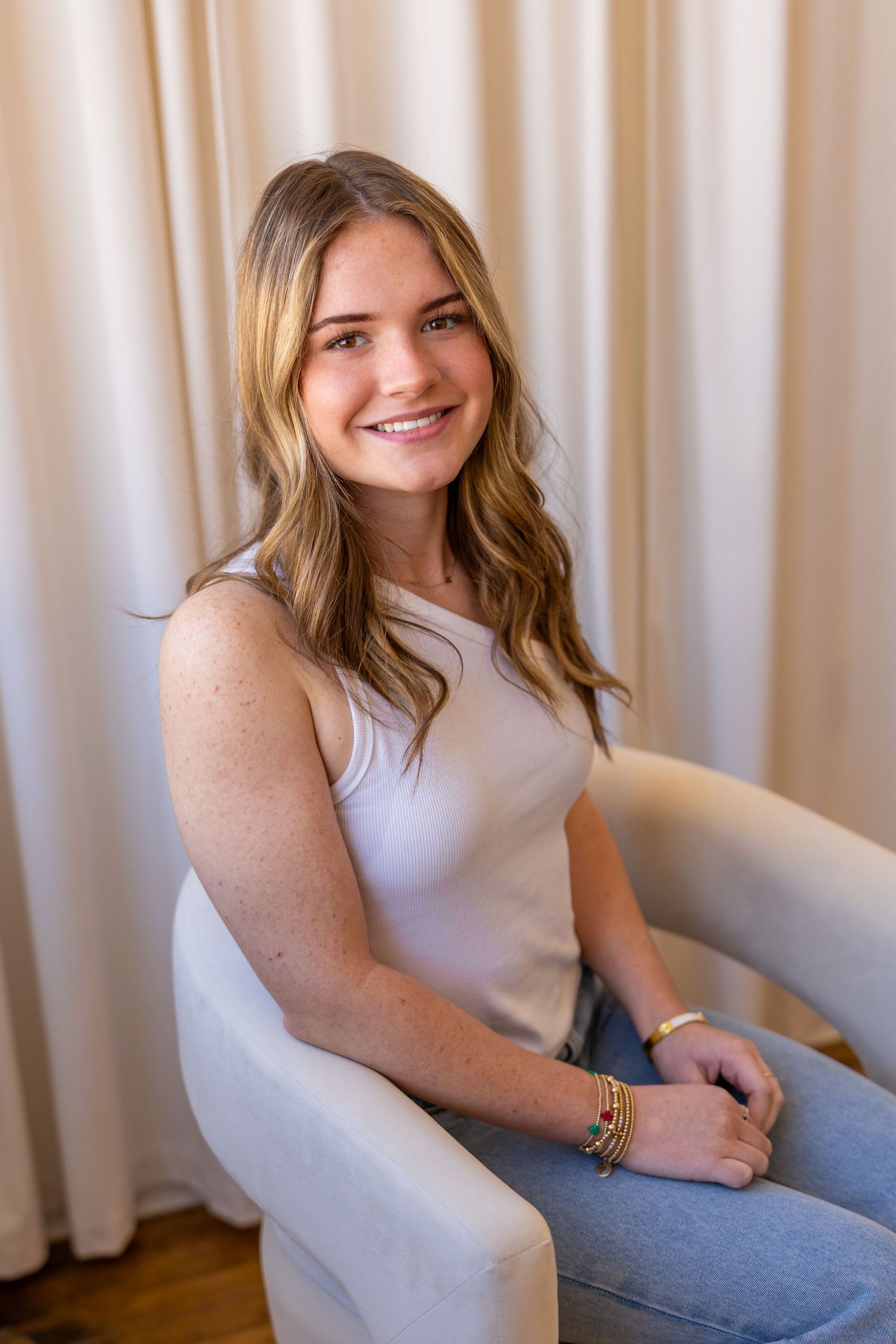 A smiling young woman with long blonde hair sitting on a chair and wearing a light colored top.