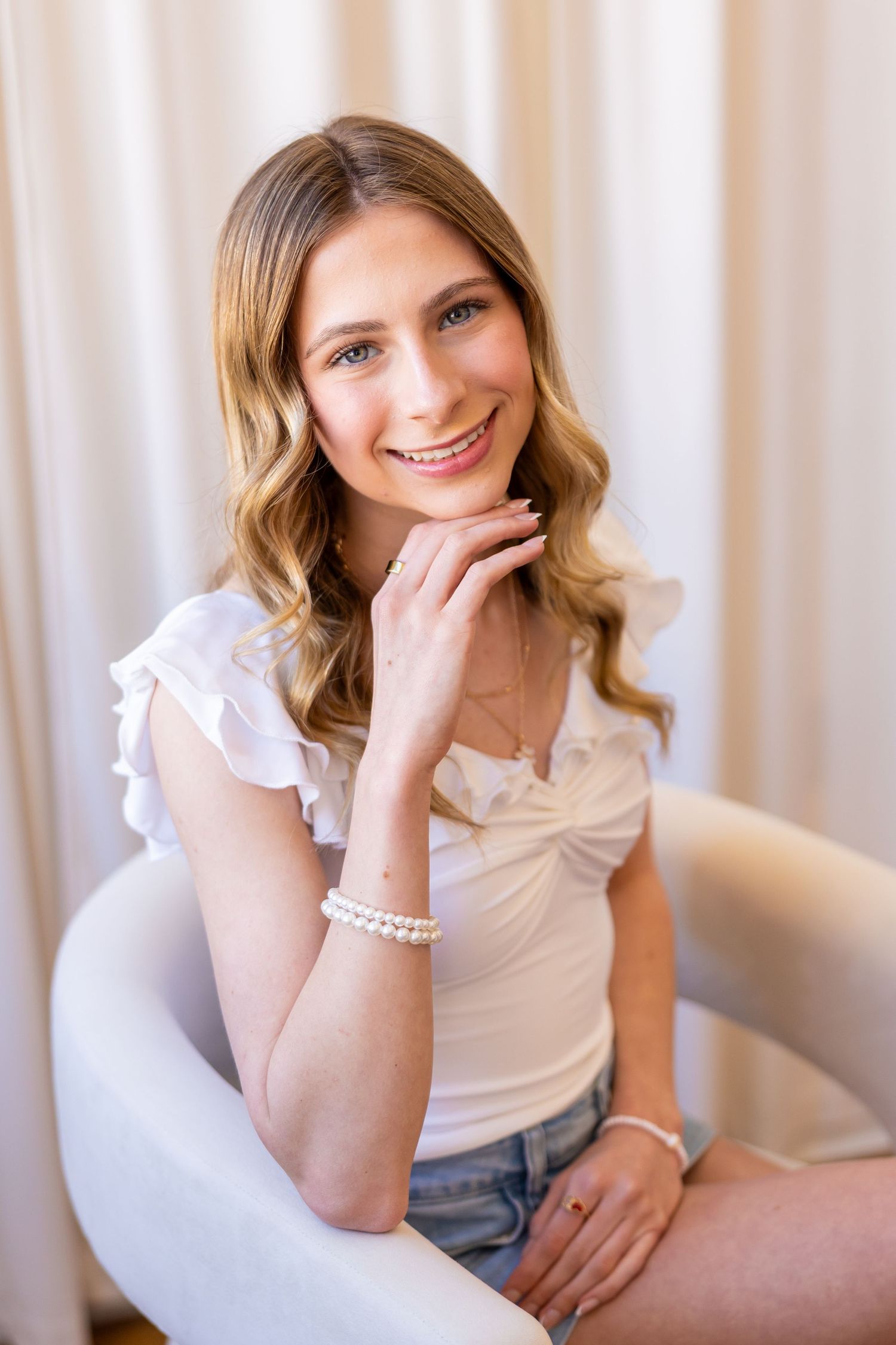 A smiling young woman with wavy blonde hair wearing a white top and sitting on a chair.