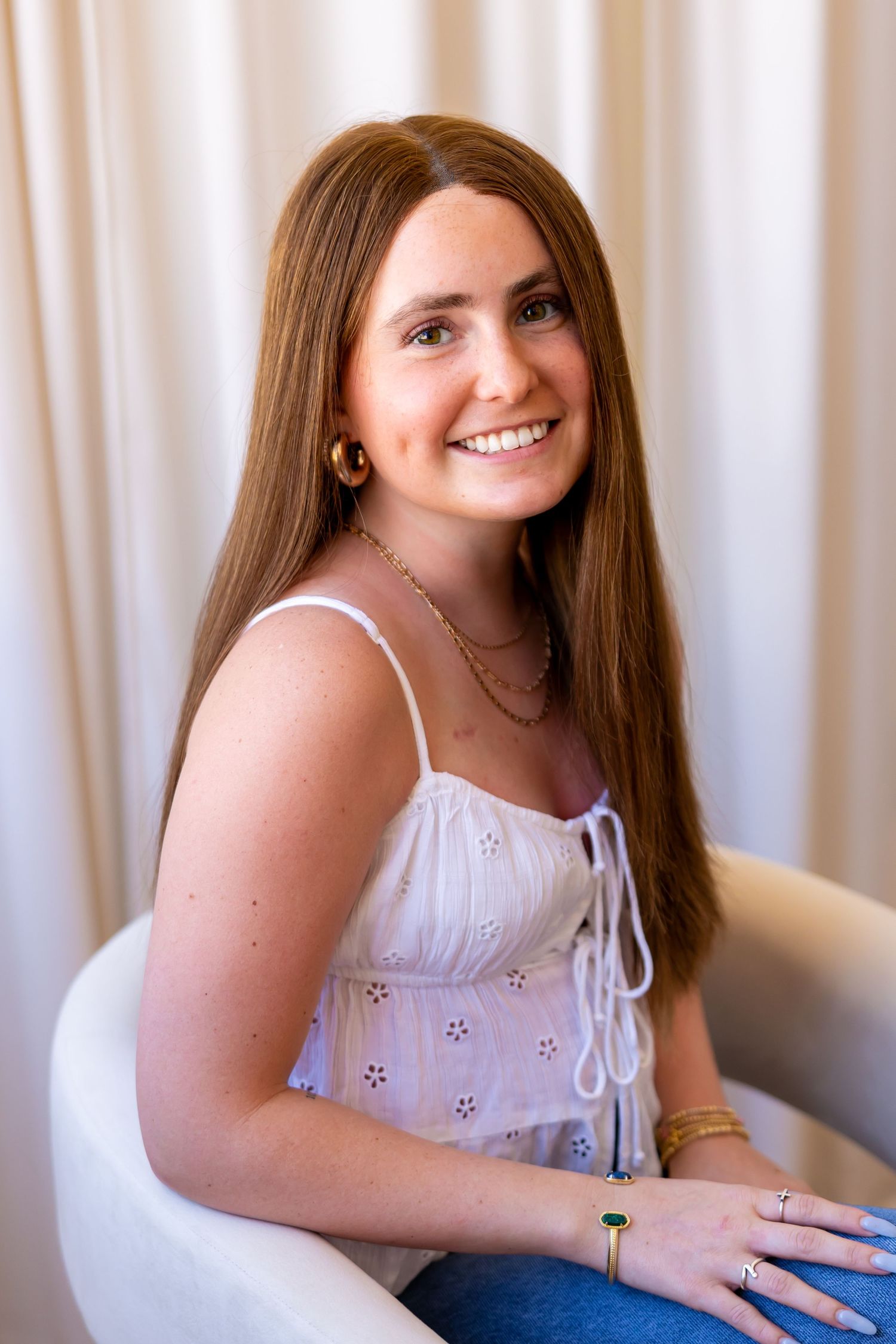 A smiling young woman with long brown hair sits in a chair against a white background.