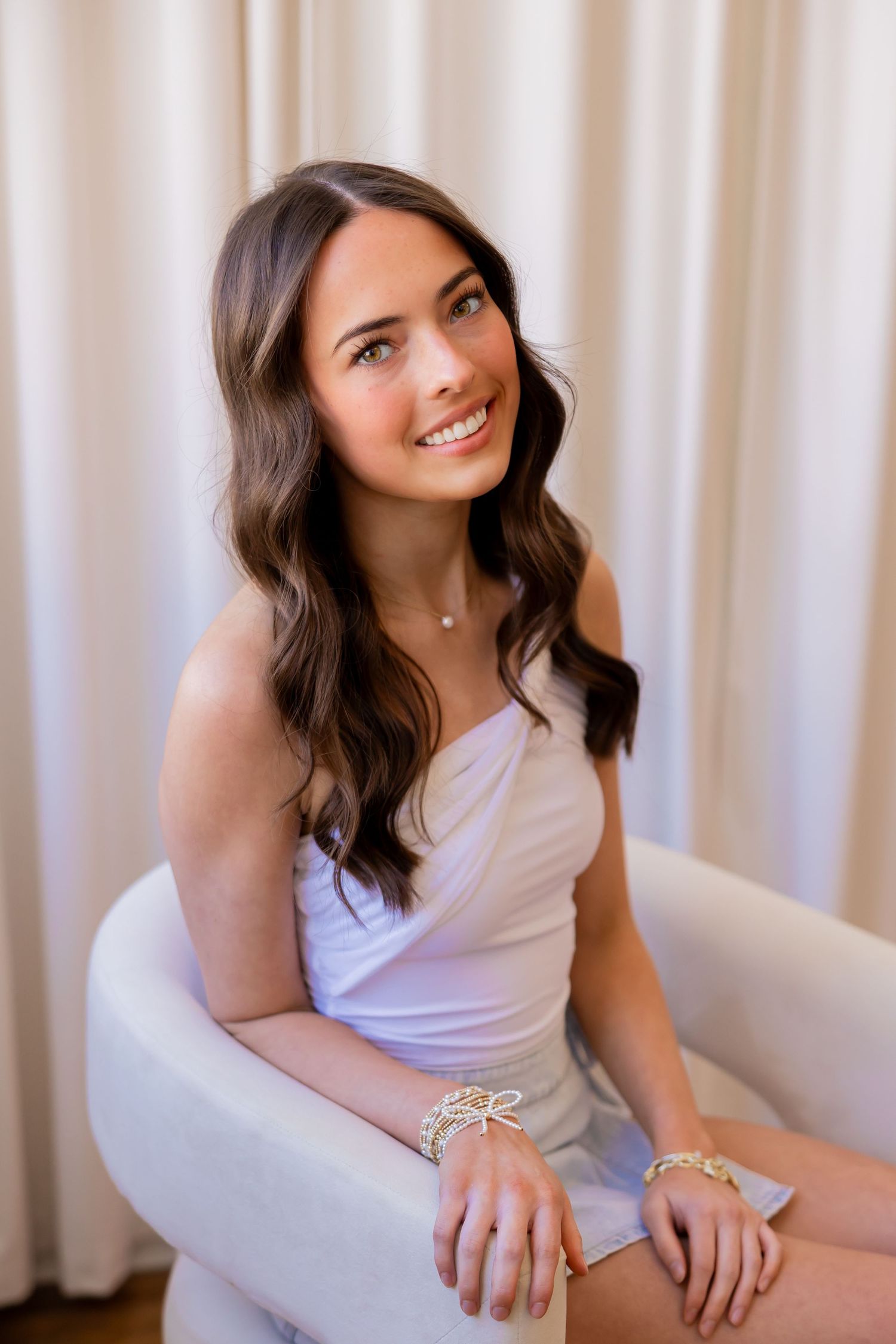 A smiling young woman with long brown hair sits in a chair against a light background.
