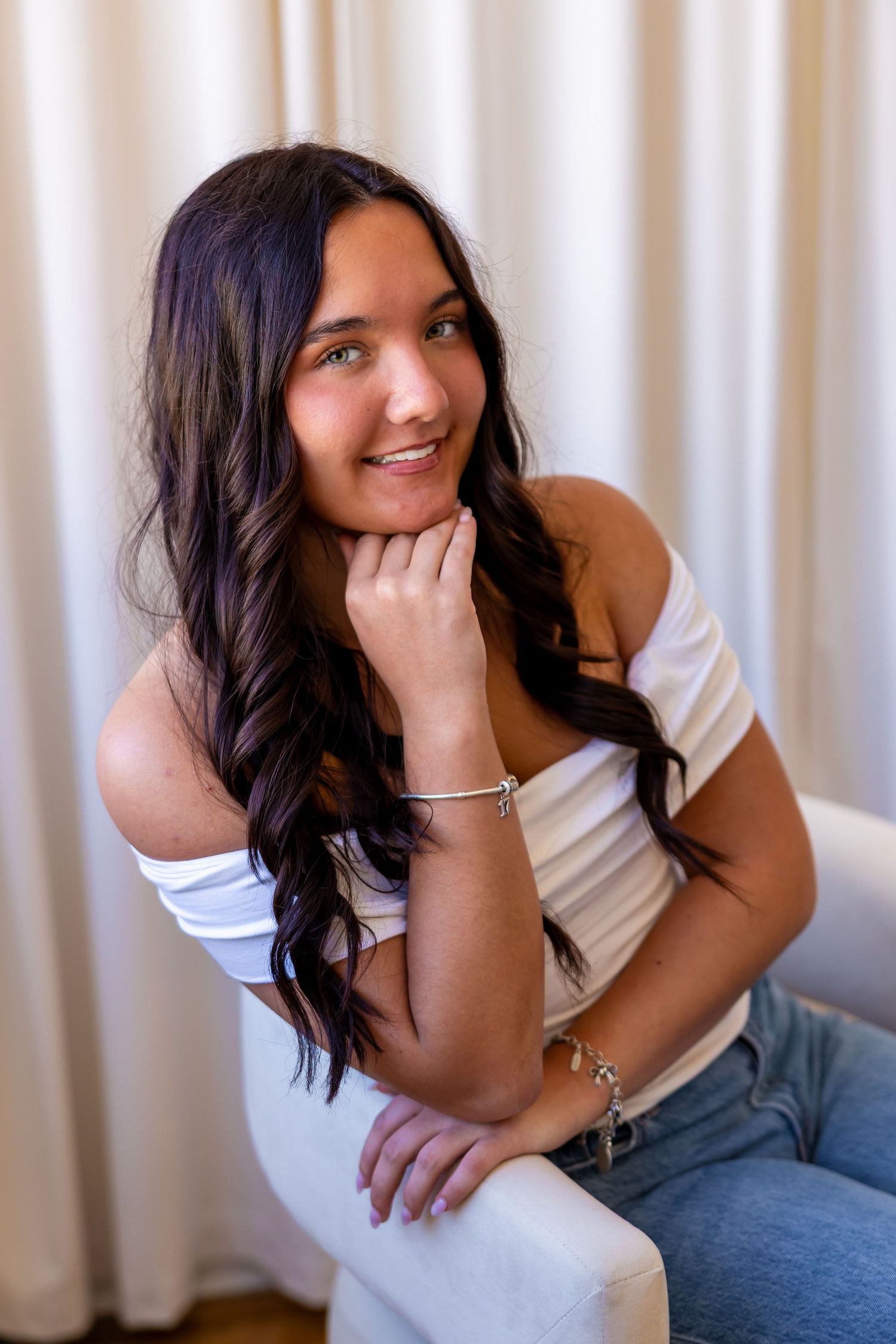 A young woman with long, dark hair smiles while sitting on a couch against a light-colored curtain backdrop.