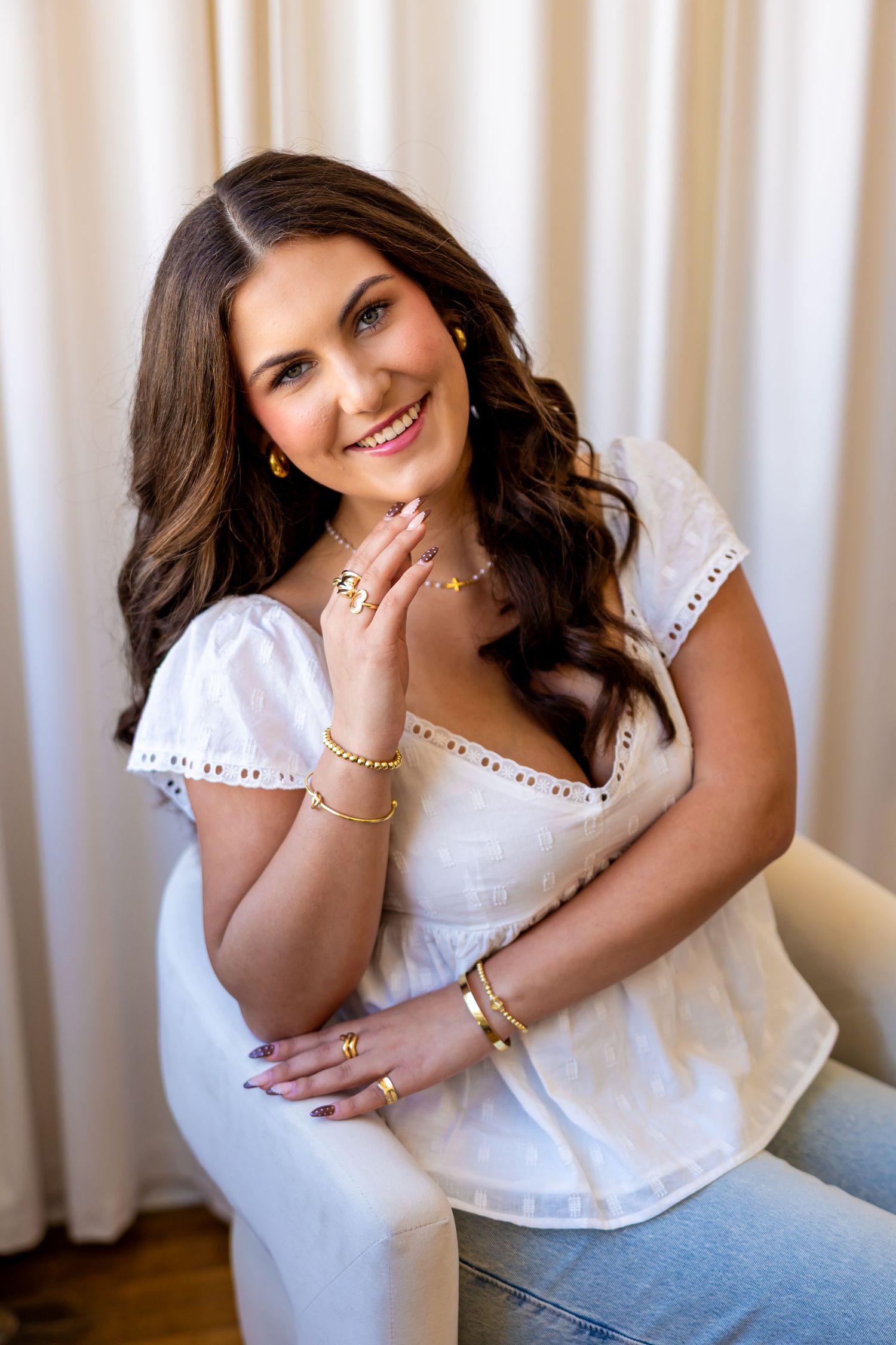 A smiling young woman with long brown hair wearing a white top sits in a chair against a light colored background.