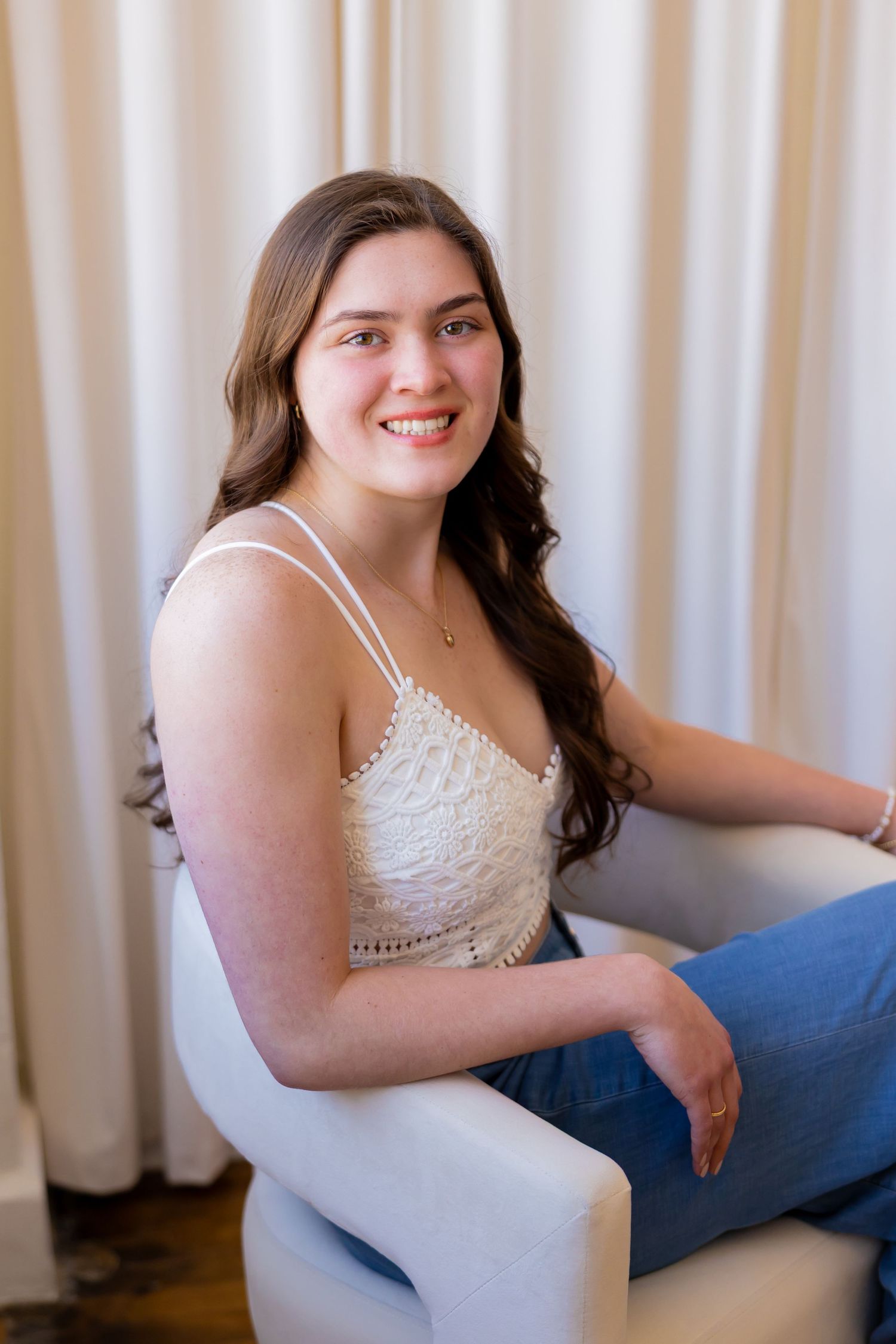 A smiling young woman with long brown hair sitting on a chair in front of a curtain.