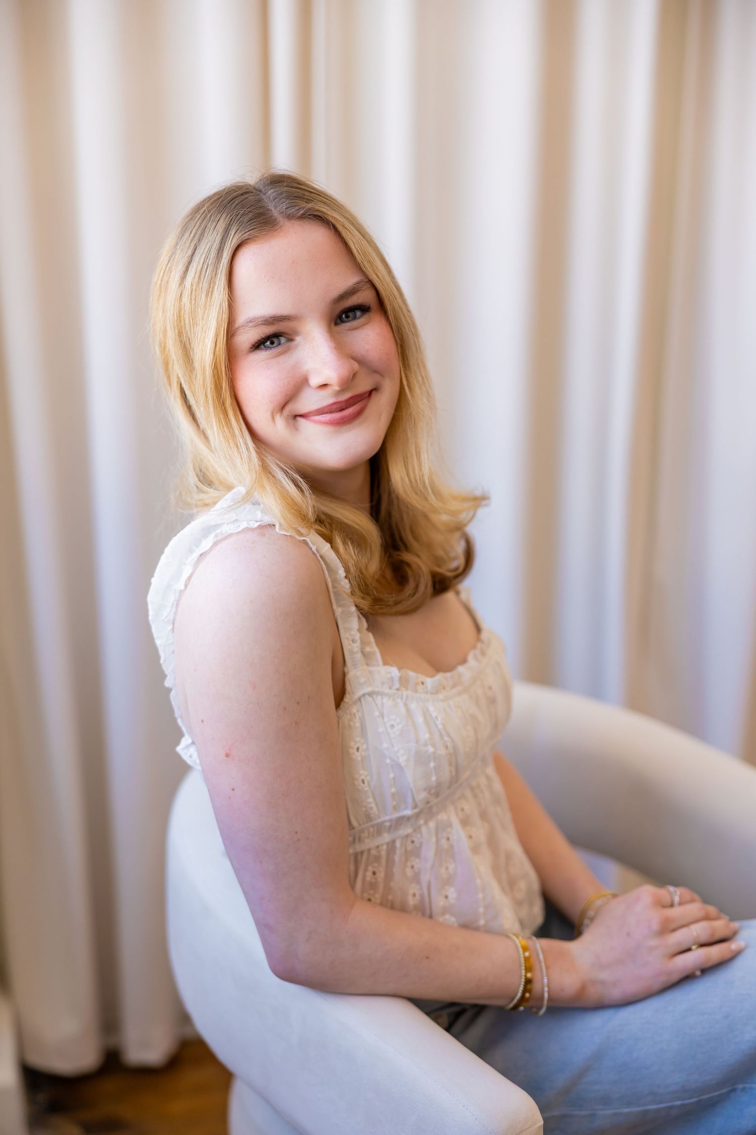 A smiling young woman with long blonde hair sitting in a chair and looking at the camera.