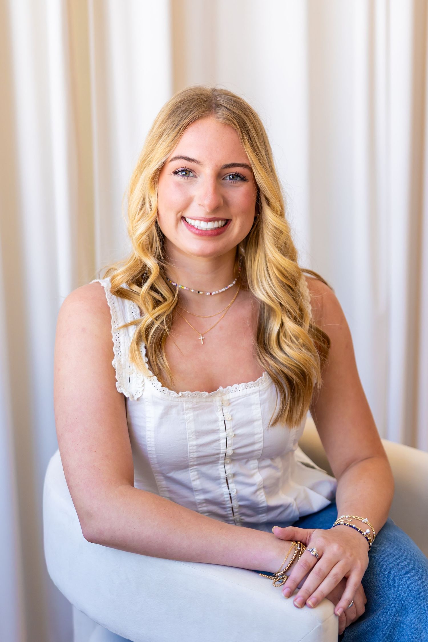 A smiling young woman with long, wavy blonde hair and a white top sits in a chair against a curtain backdrop.