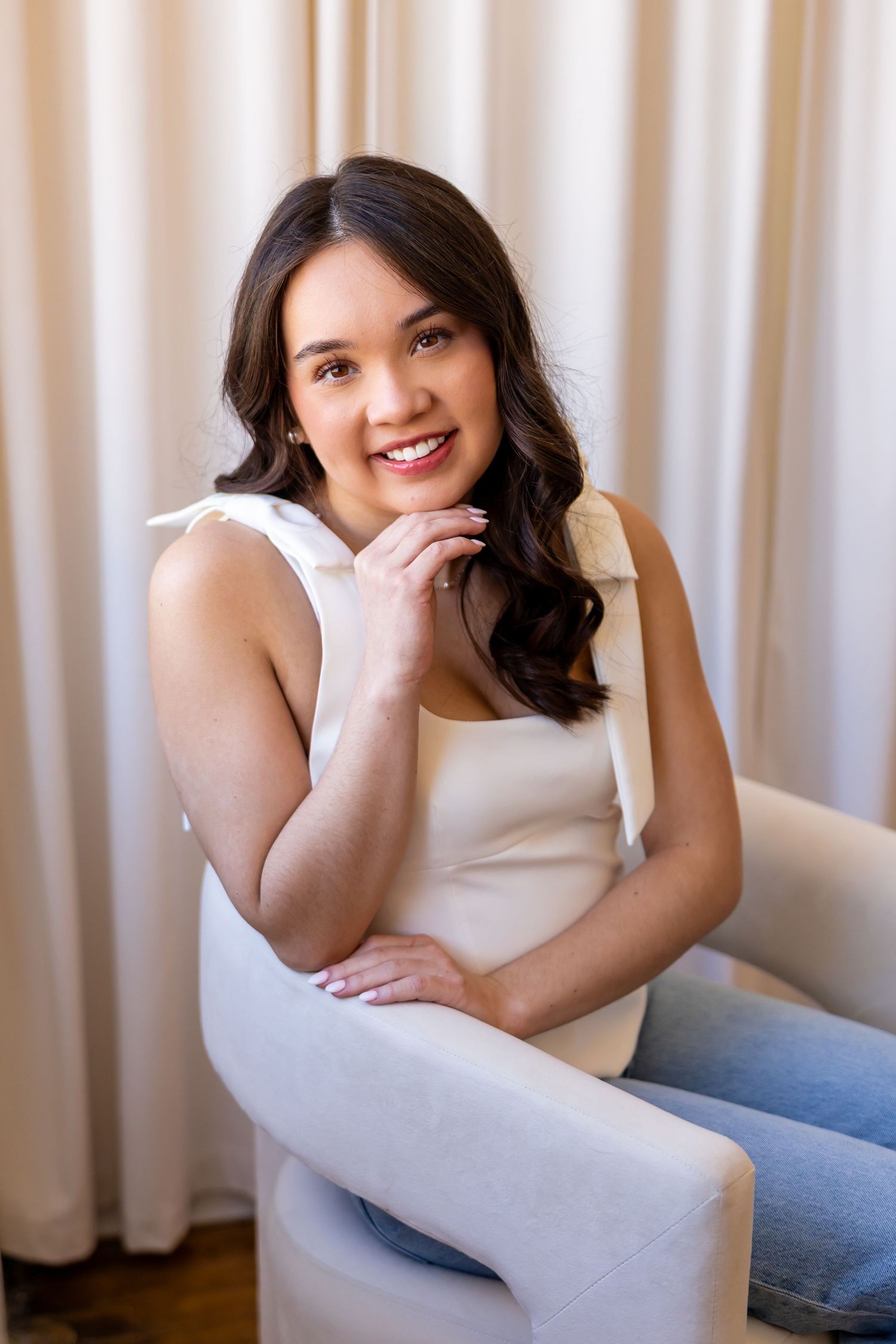 A smiling young woman with long dark hair sitting in a chair and looking at the camera.