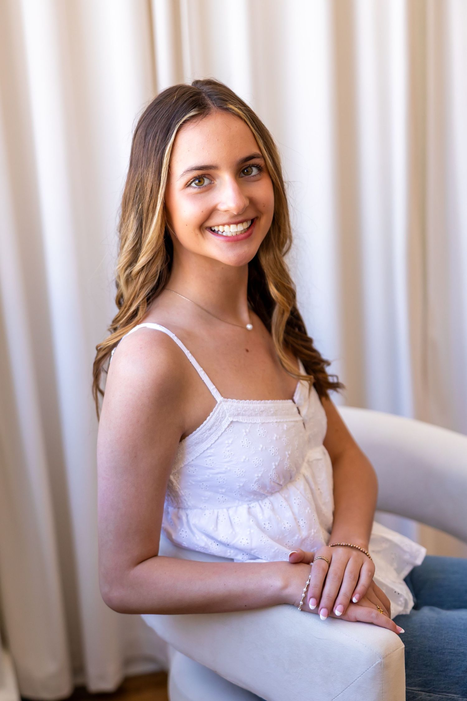 A smiling young woman with long brown hair sitting on a couch and looking directly at the camera.