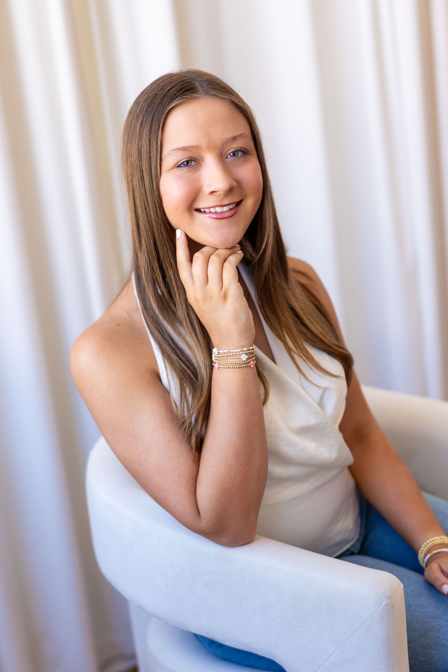A smiling young woman with long brown hair sitting in a chair against a light background.