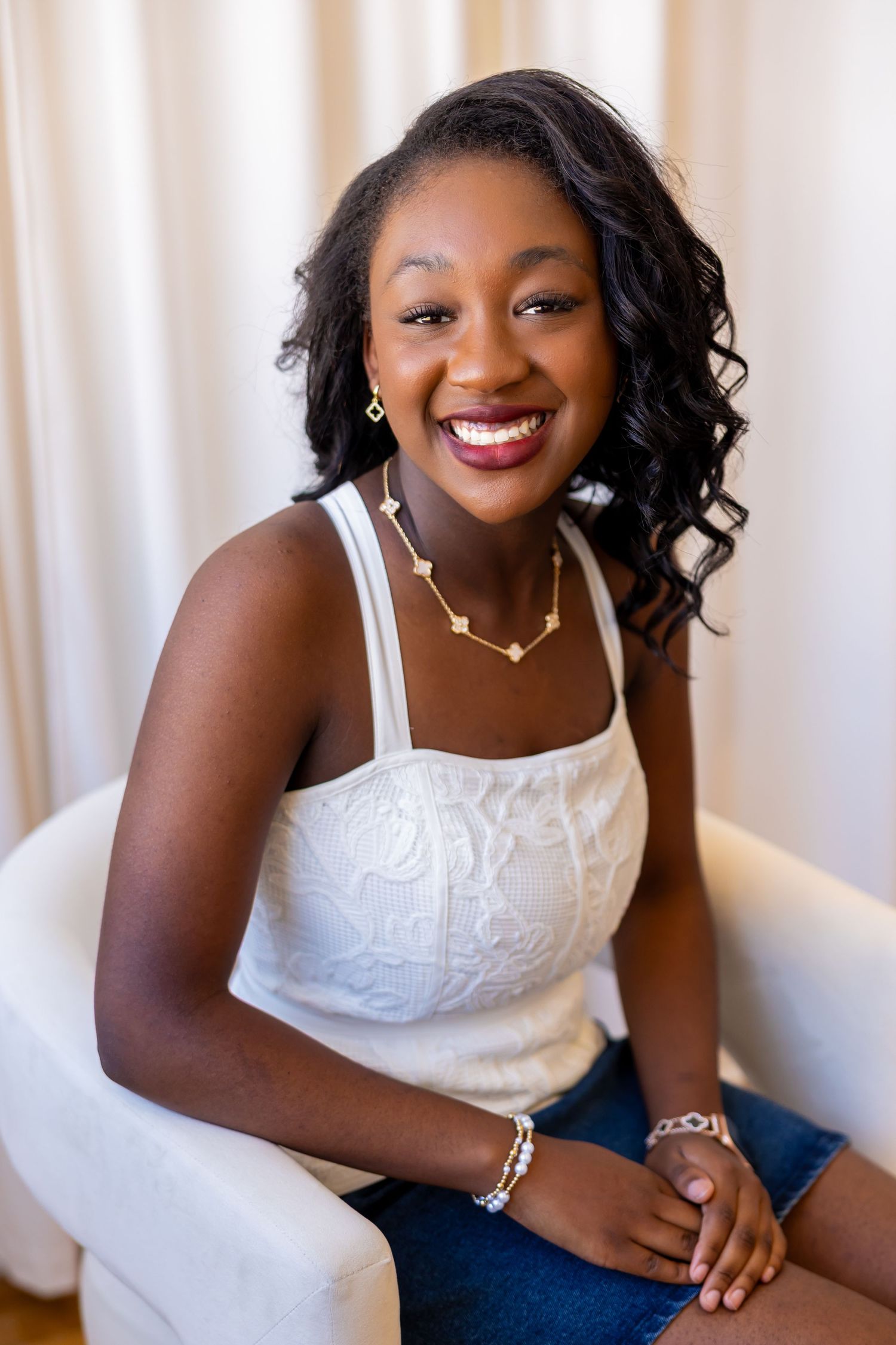 A smiling young woman with curly dark hair wearing a white tank top and sitting on a chair.