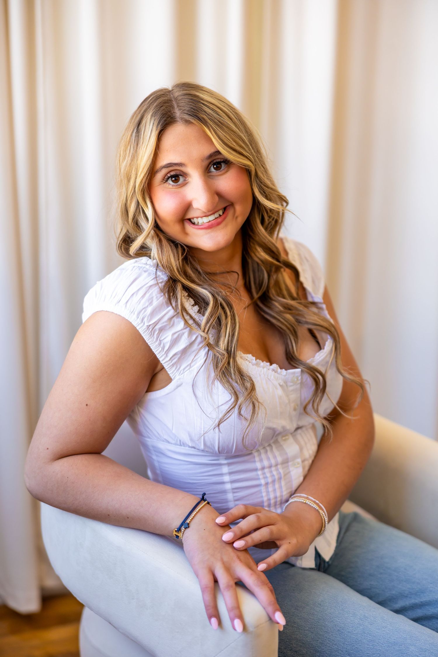 A young woman with long blonde curly hair smiles while sitting on a couch in a room with curtains.