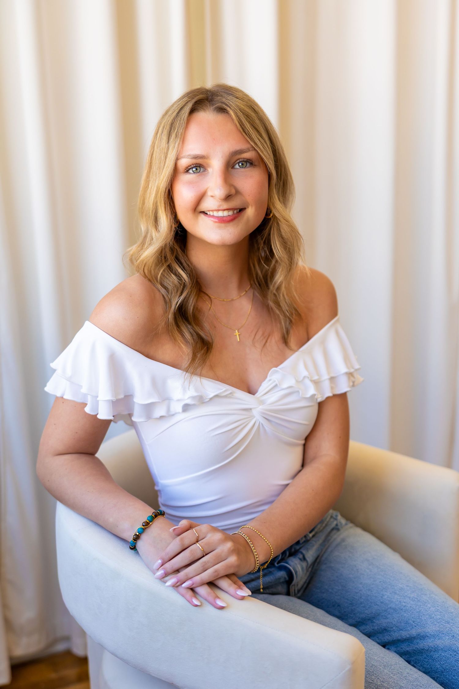 A smiling young woman with wavy blonde hair sits on a chair wearing a white off-the-shoulder top and jeans.