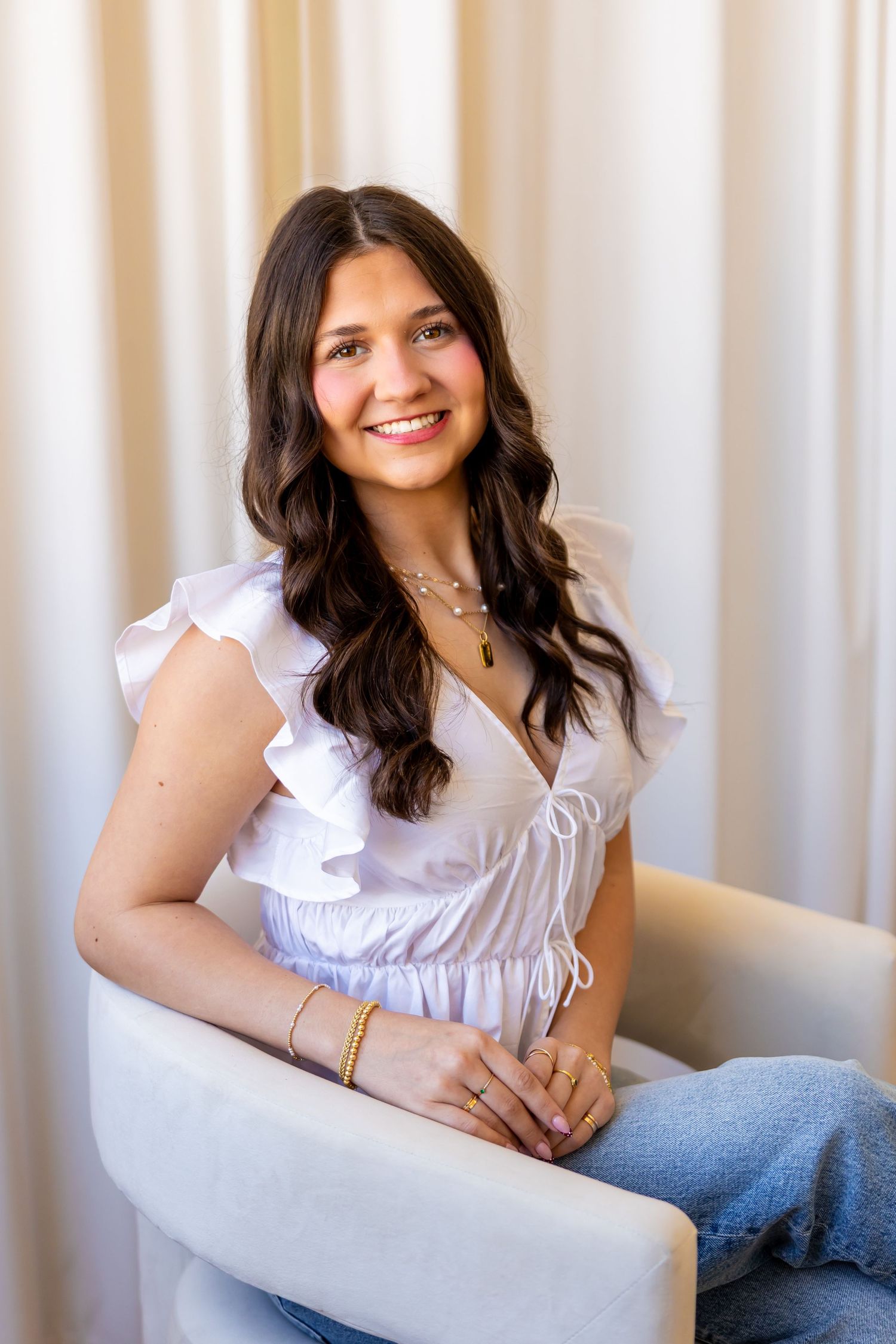 A smiling young woman with long dark hair wearing a white ruffled top and sitting on a couch.