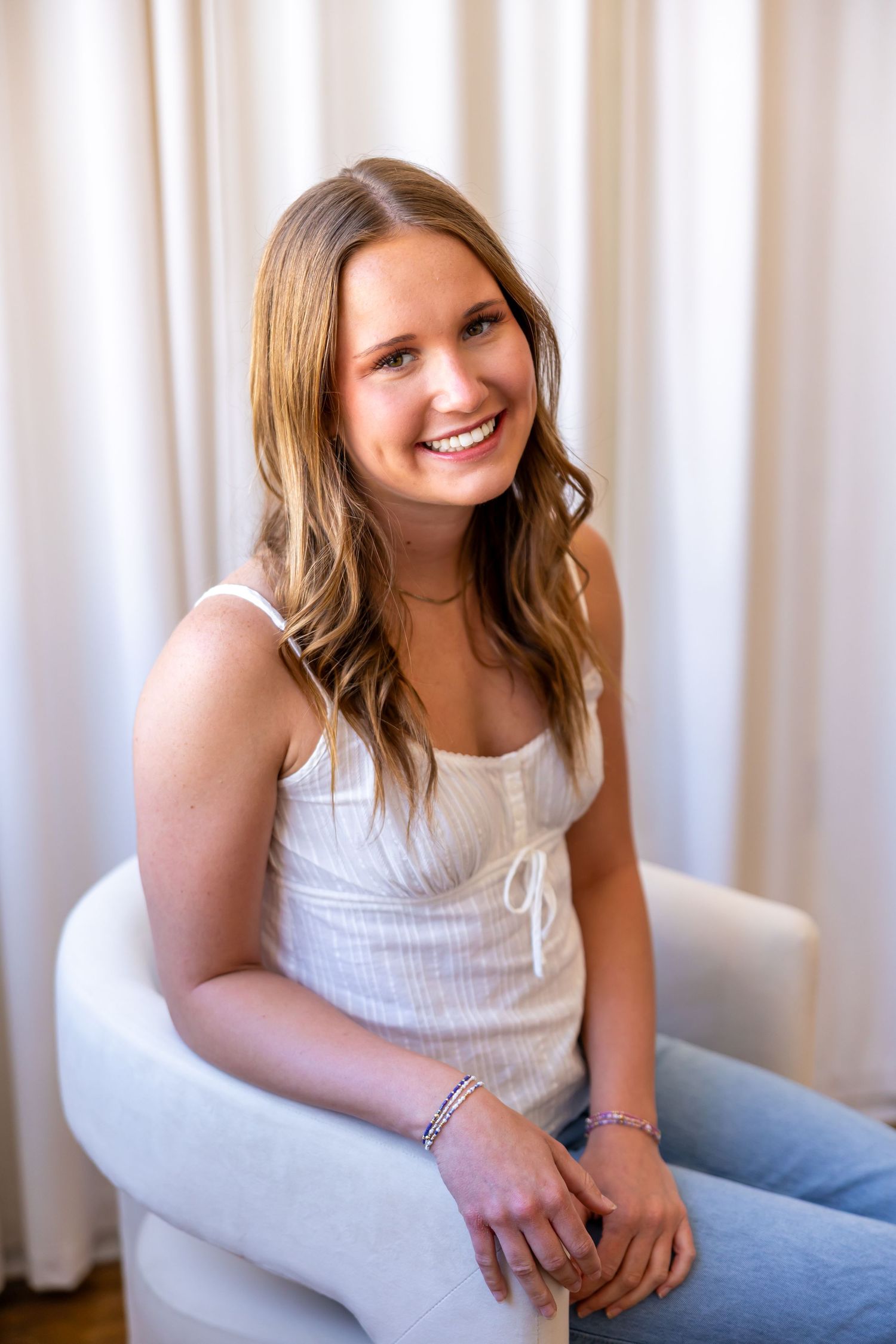 A smiling young woman with long blonde hair wearing a white tank top and sitting in a chair.