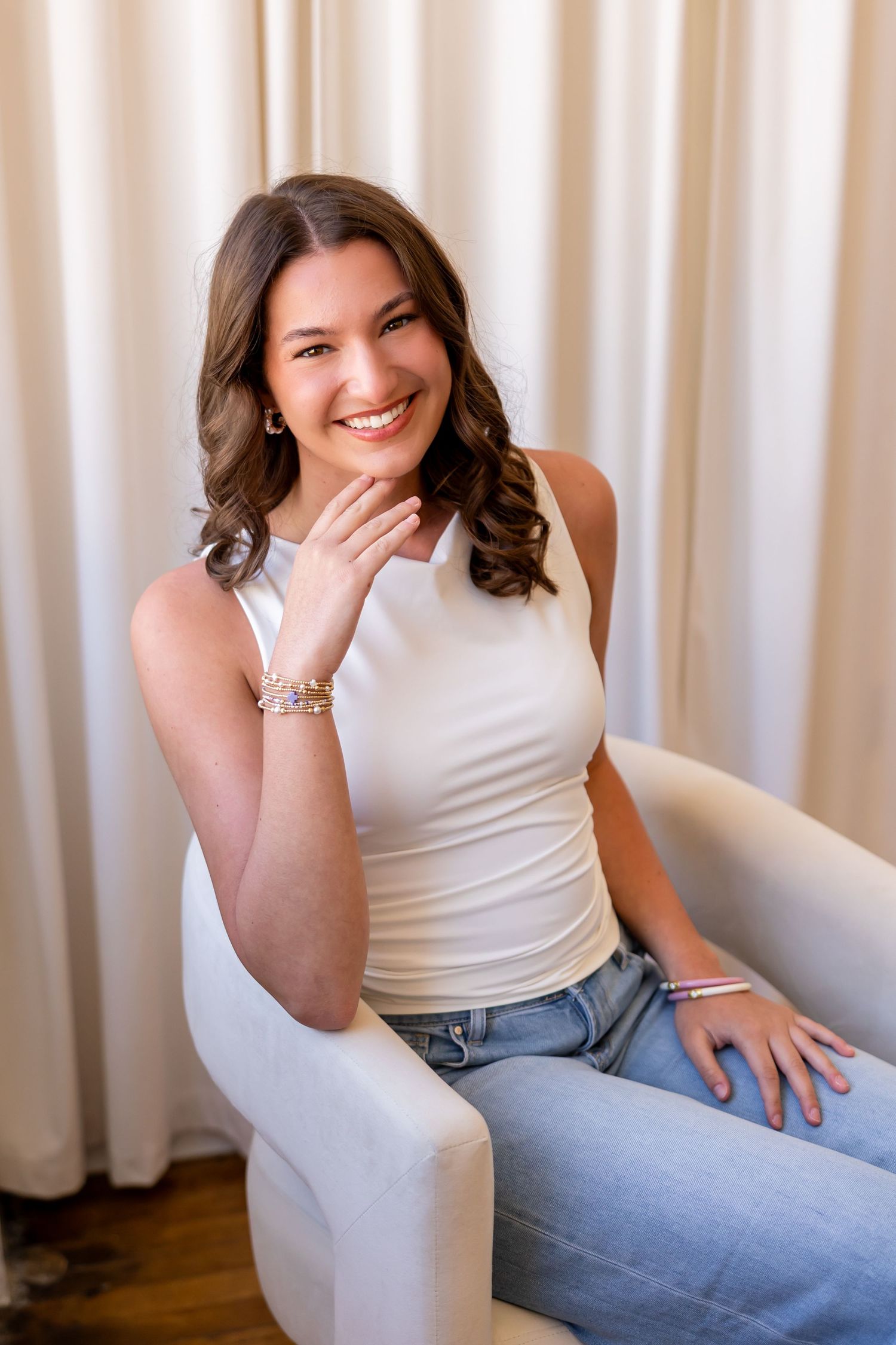 A young woman with long brown curly hair smiles warmly while sitting on a chair.