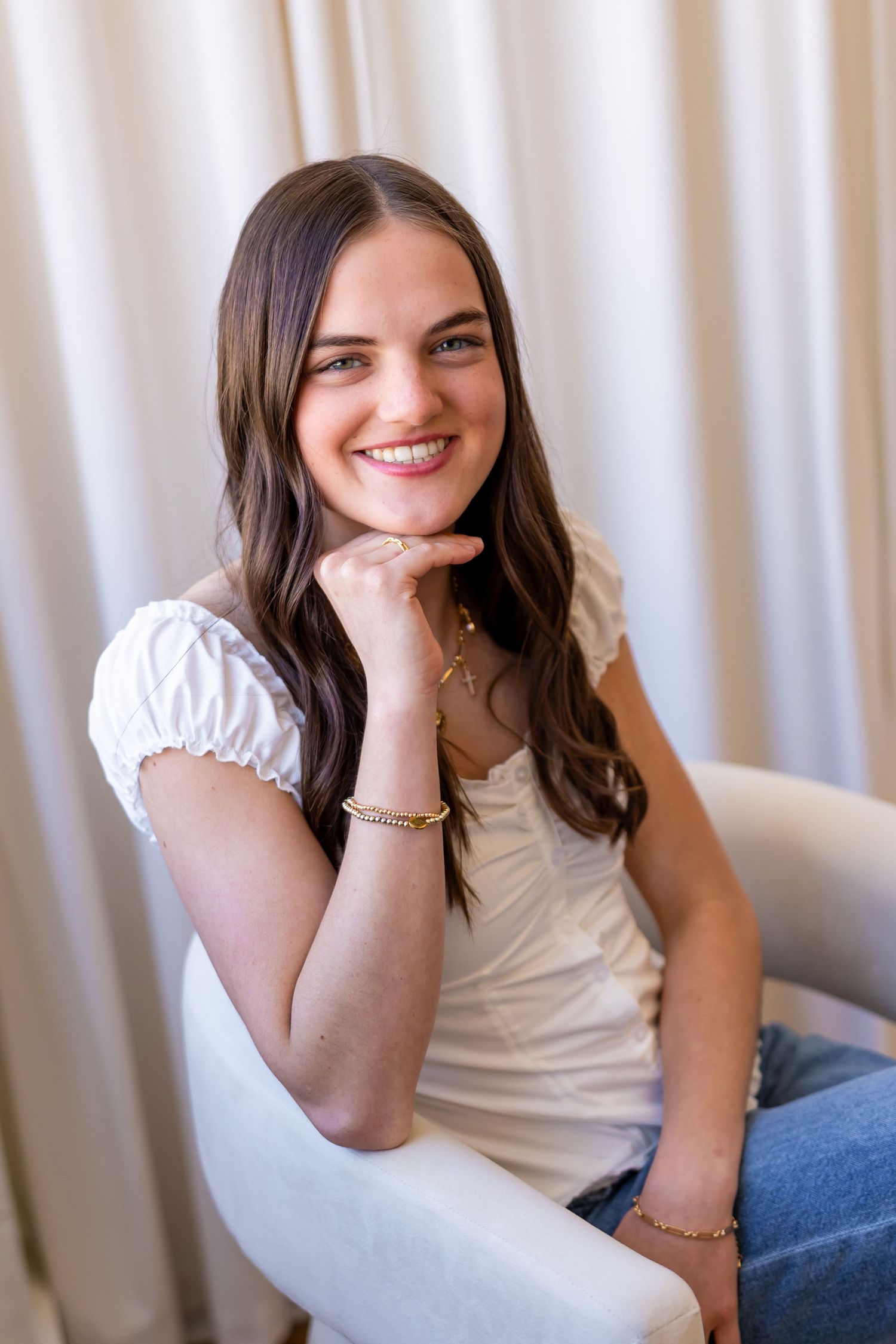 A smiling young woman with long dark hair sits in a chair against a light-colored curtain background.