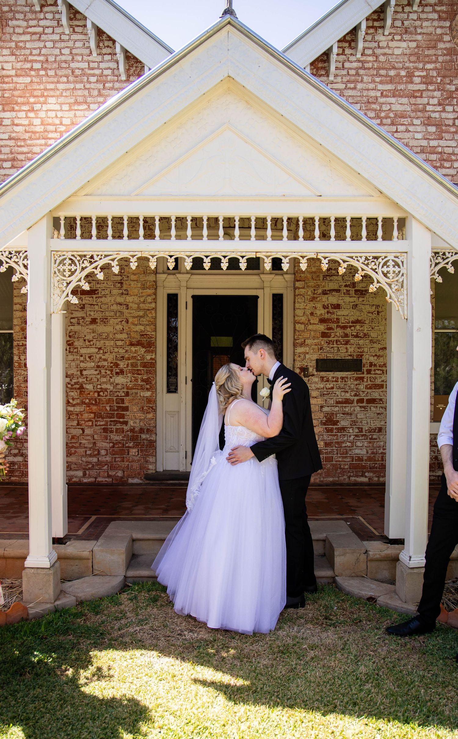 A couple in formal wedding attire share a moment on a brick building's porch with ornate Victorian-style trim details.