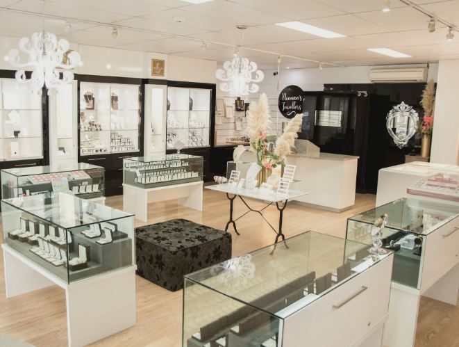 A modern jewelry store interior with glass display cases, white chandeliers, and hardwood flooring illuminated by bright lighting.