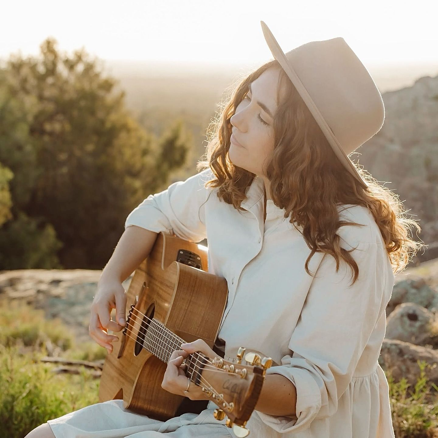 A musician in a flowing white dress plays acoustic guitar in golden sunlight among desert hills at sunset.