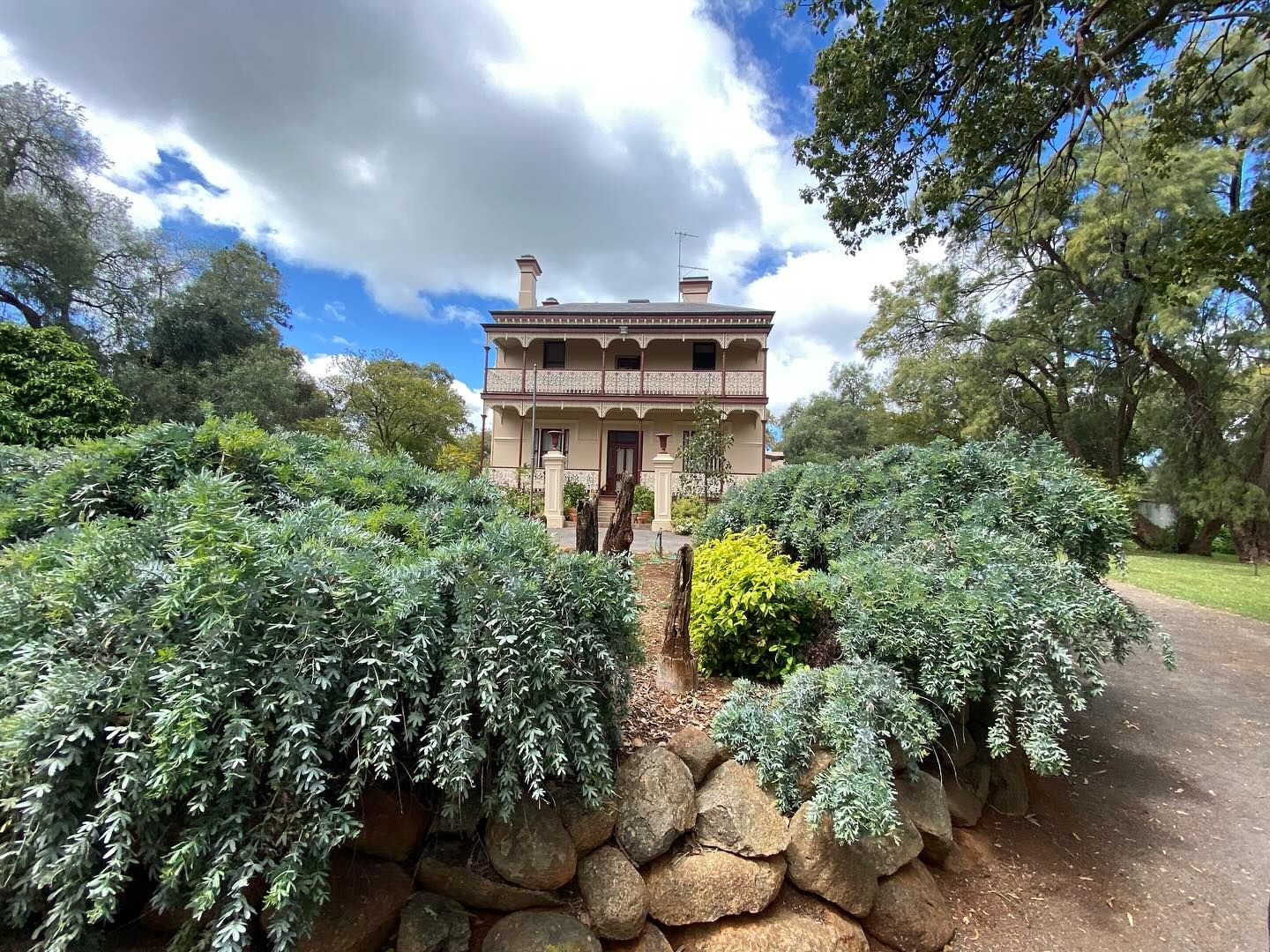 Historic stone mansion with wraparound porch stands behind lush garden beds on a sunny day in Australia.