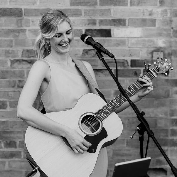 A musician performs with an acoustic guitar and microphone against a rustic brick wall in black and white.