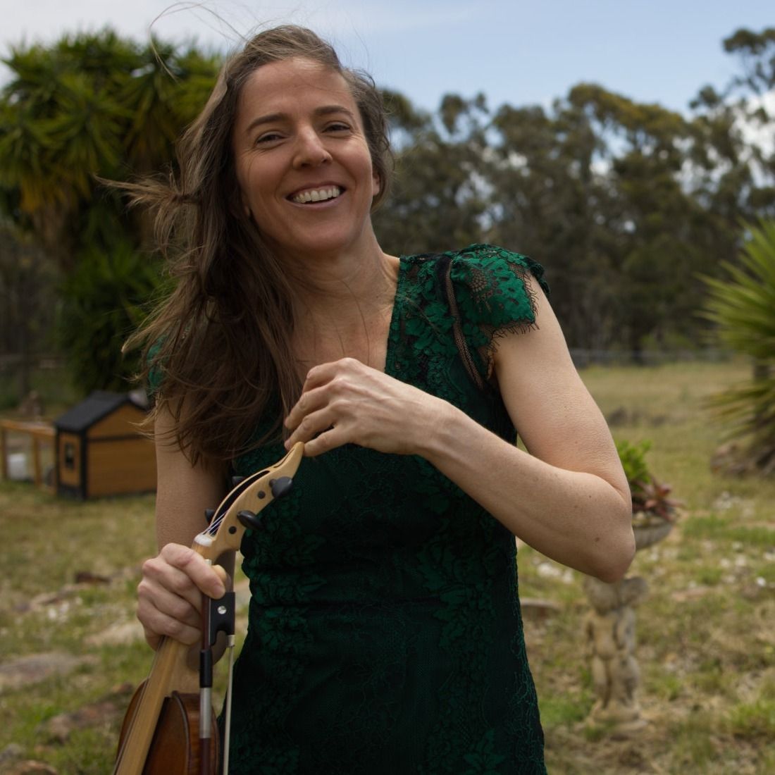 Someone in an emerald green dress smiles joyfully outdoors with windswept hair against a natural landscape backdrop.