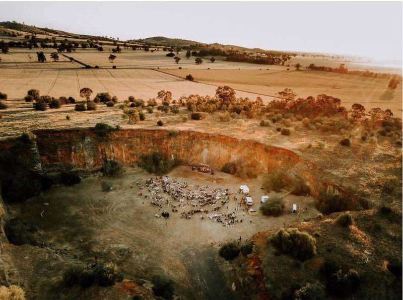 An aerial view shows a herd of sheep grazing in a rustic Australian outback landscape with scattered eucalyptus trees.
