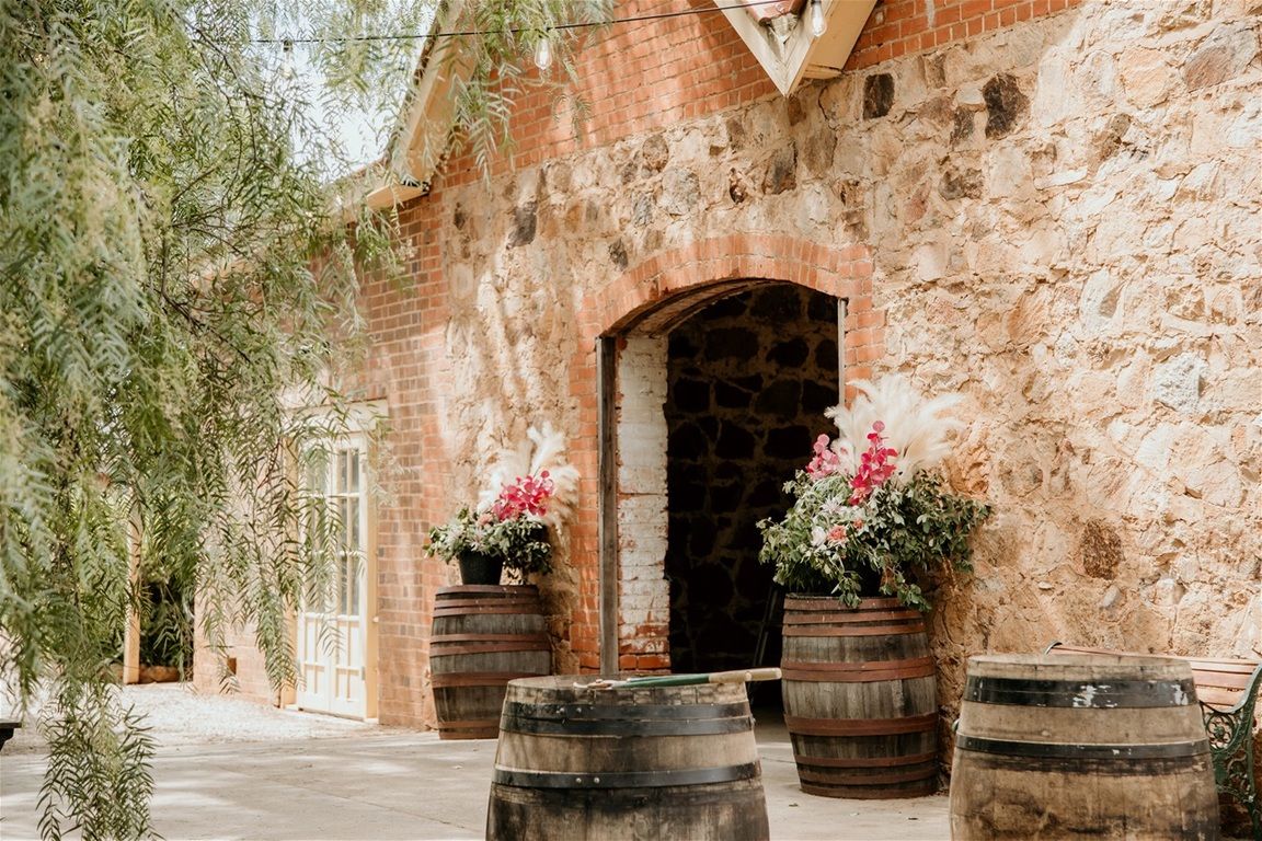 Rustic stone winery entrance with wooden barrels and pink flowers in bloom outside arched wooden doorway.