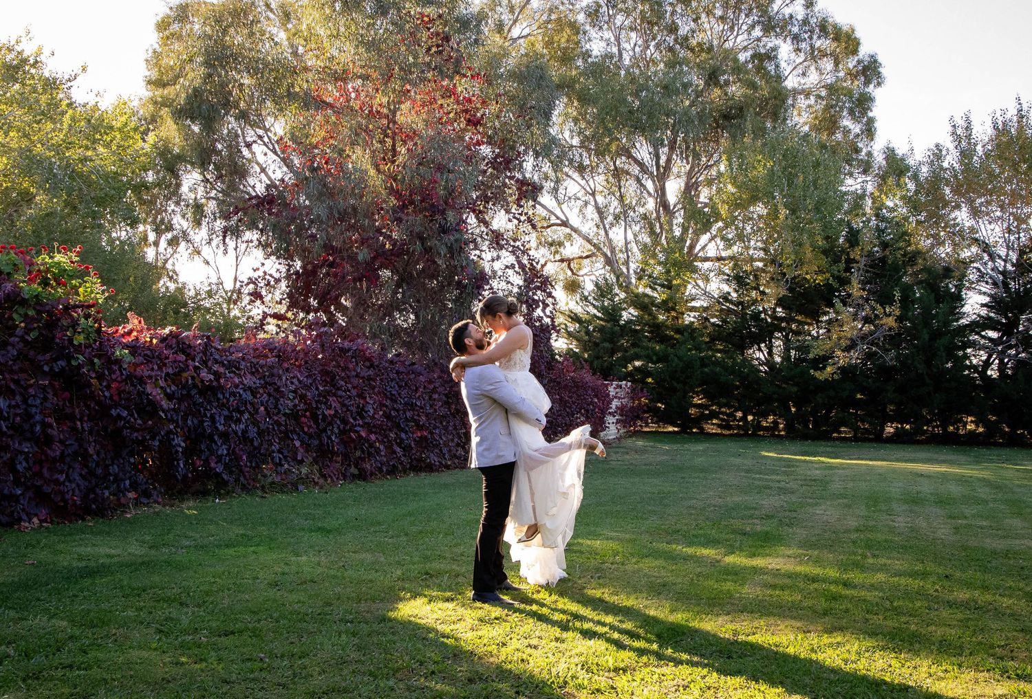 A couple shares a romantic embrace while dancing on a sunlit lawn surrounded by lush garden hedges and trees.