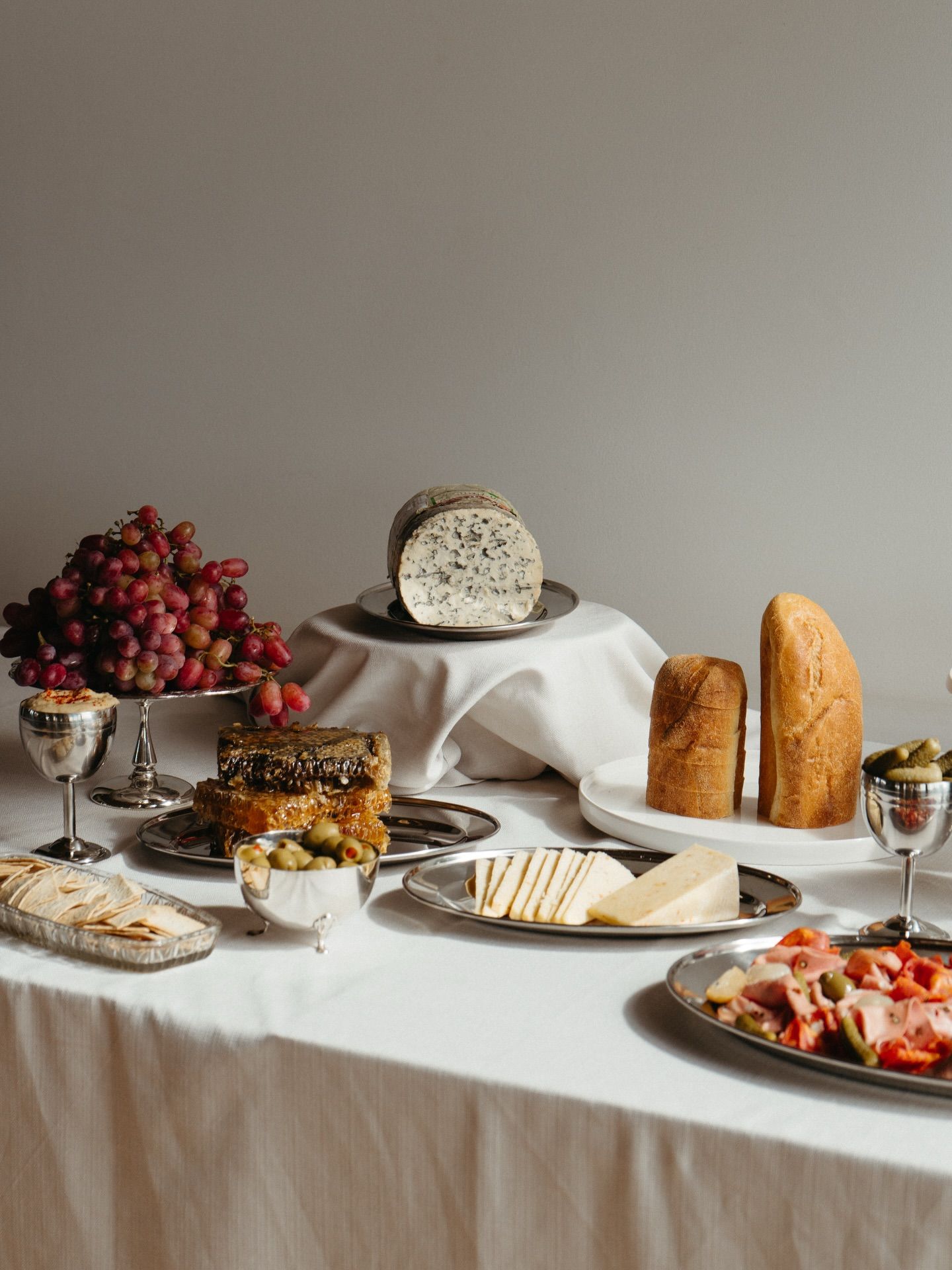 Elegant charcuterie spread with blue cheese, grapes, bread, and meats on silver platters atop a white linen table.