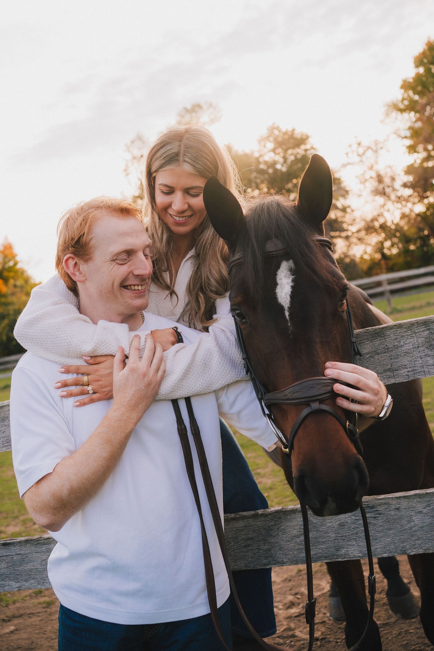 A couple shares joyful moments with a dark brown horse by a wooden fence during golden hour sunset.