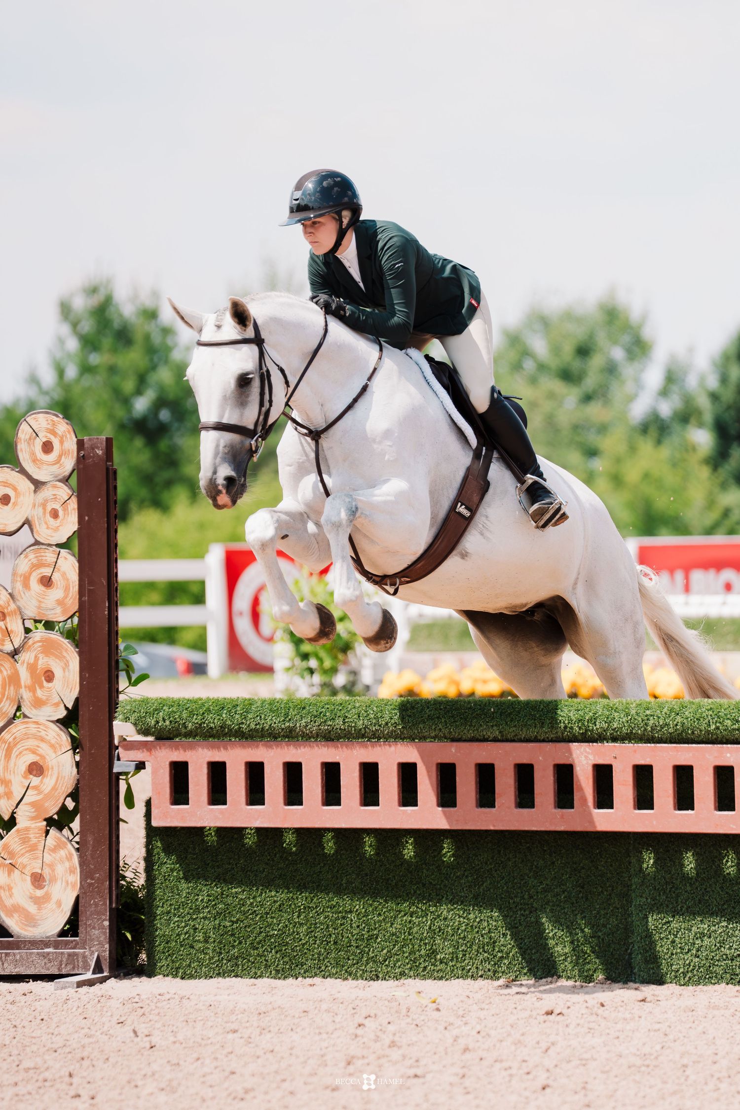 White horse and rider clearing a wooden show jumping fence during an equestrian competition on a sandy arena.