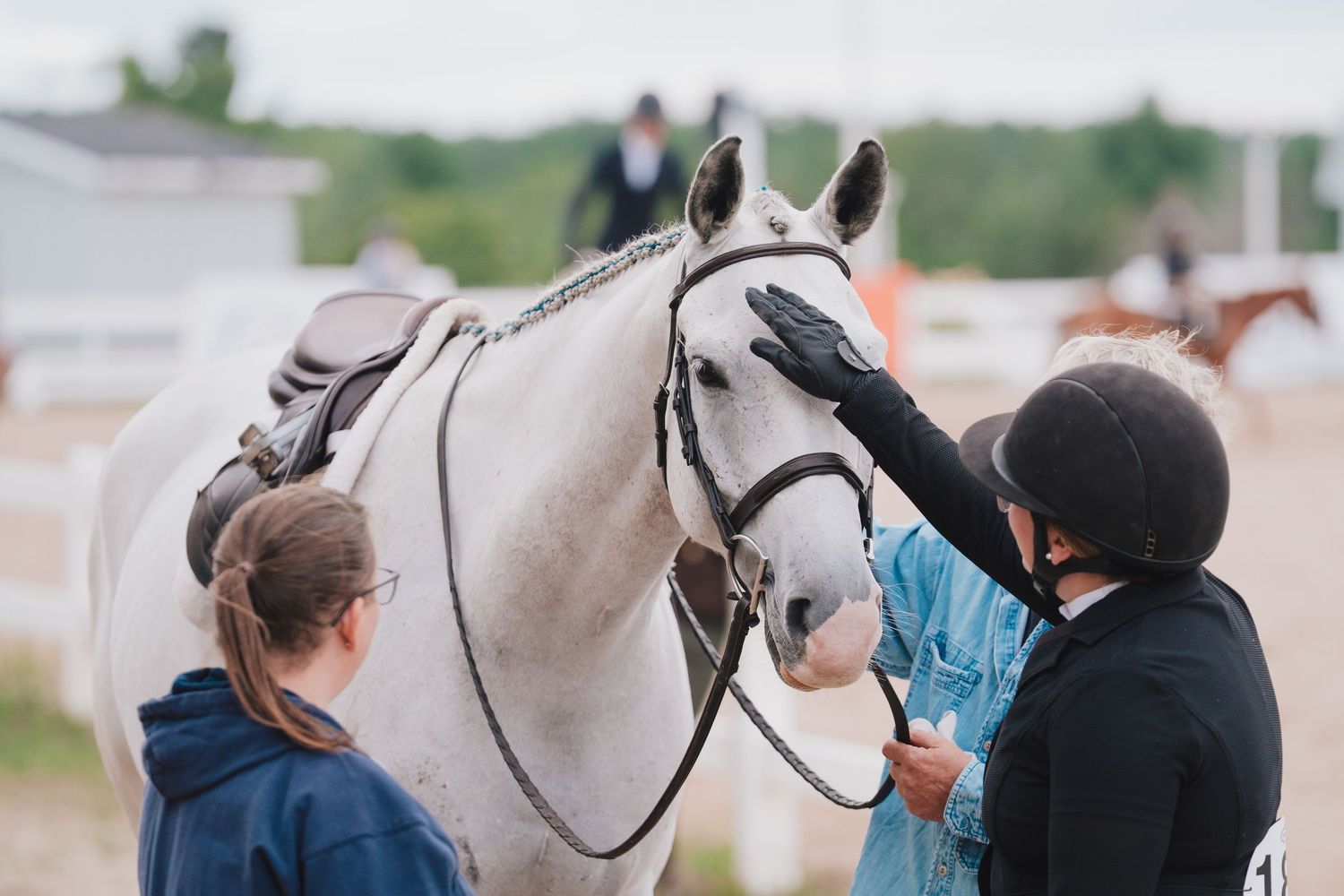 Two people in riding gear tend to a beautiful white horse wearing show tack at an equestrian event.