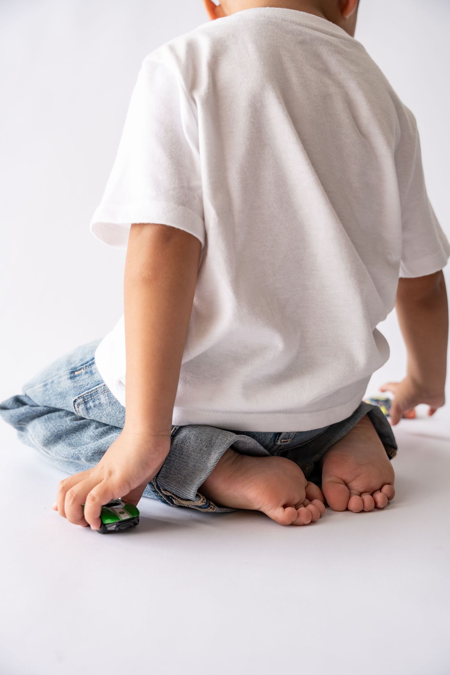 Young child sitting on white floor playing with a small toy car, wearing white t-shirt and jeans, viewed from behind.