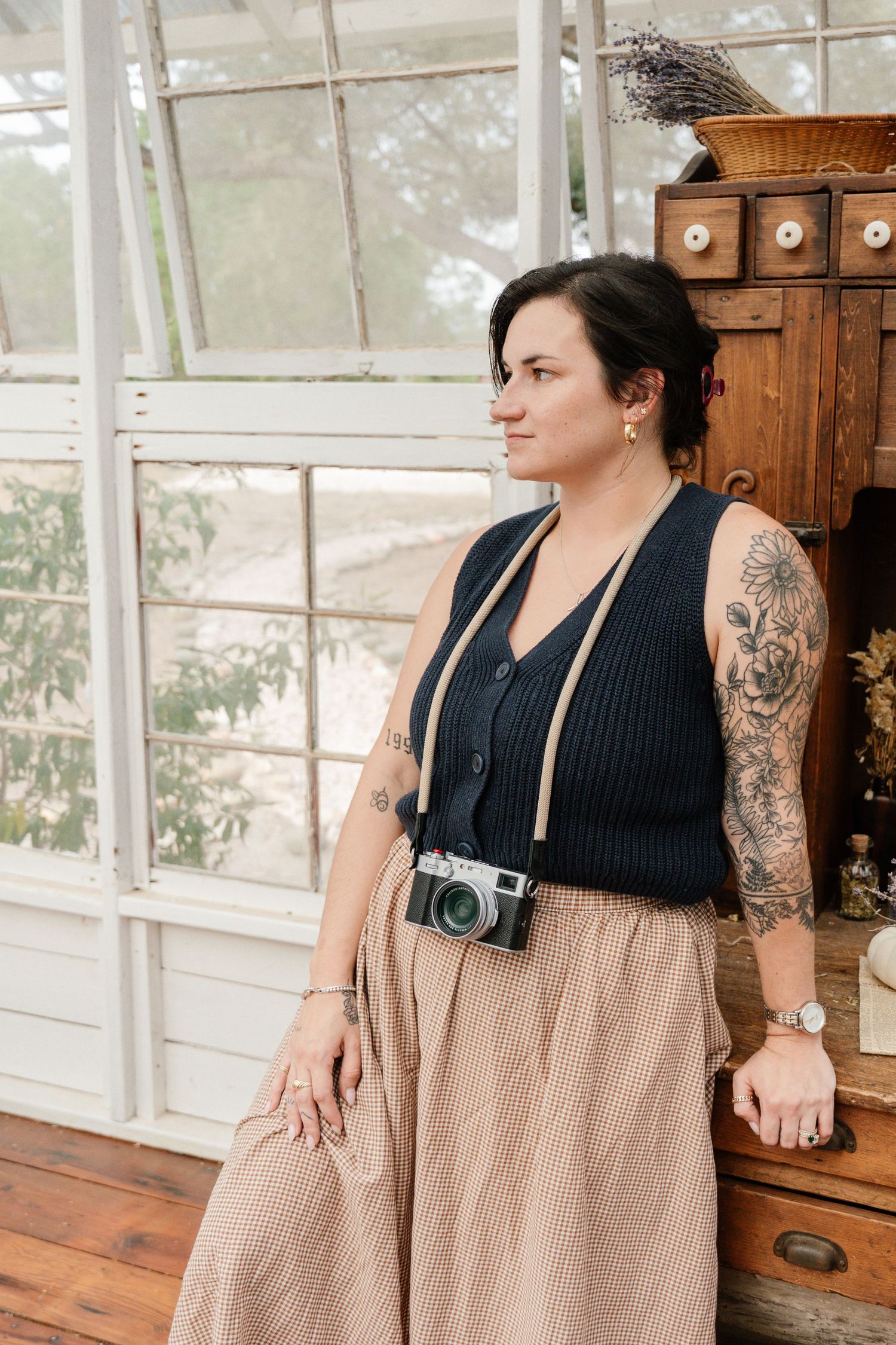 Woman with tattoos wearing a black vest and beige skirt holds a vintage film camera in a rustic greenhouse setting.