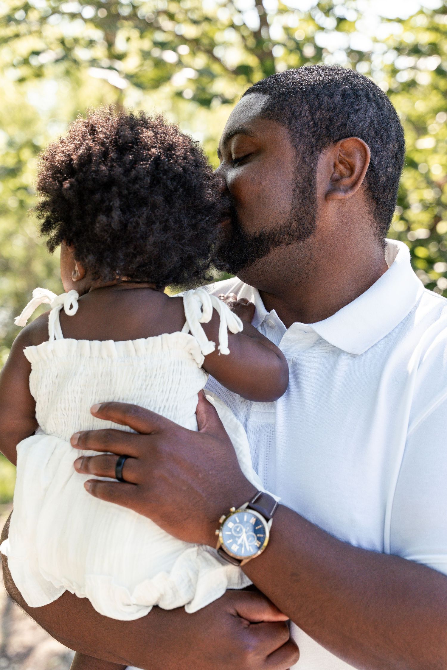 A loving Black father kisses his young daughter on the cheek while holding her in a sunny outdoor setting.
