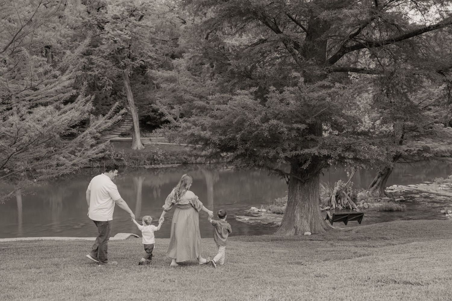 Black and white photo of a family holding hands walking in a park near a pond with large trees and a swing.
