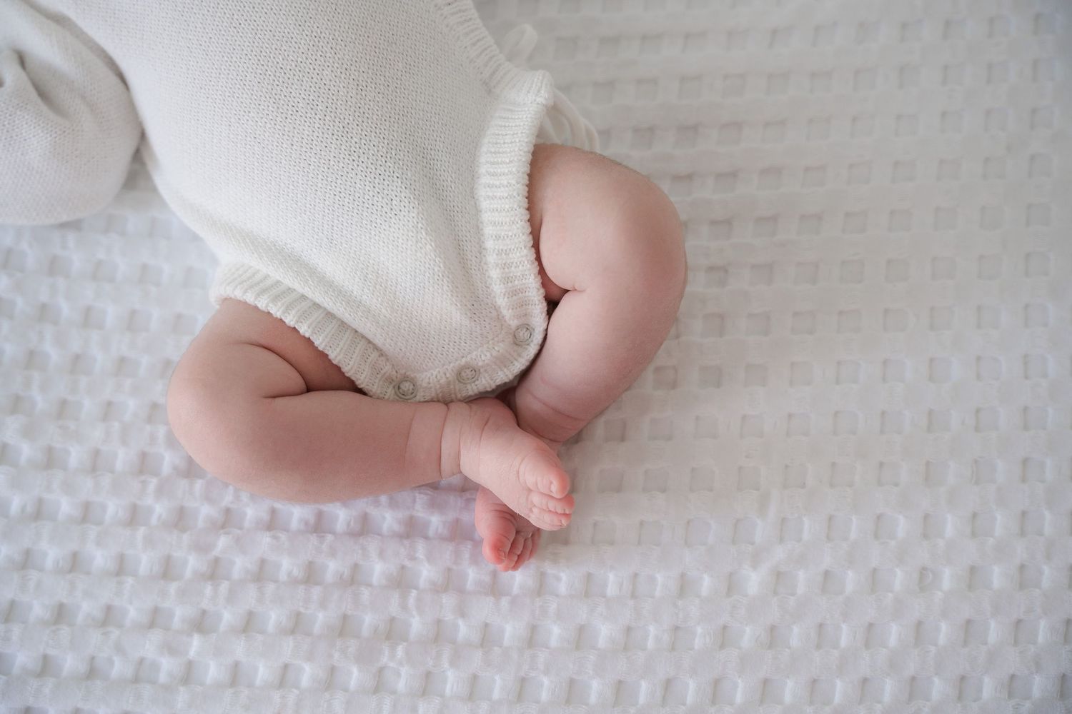Close-up of a sleeping newborn baby in a white knit outfit lying on a soft white textured blanket.