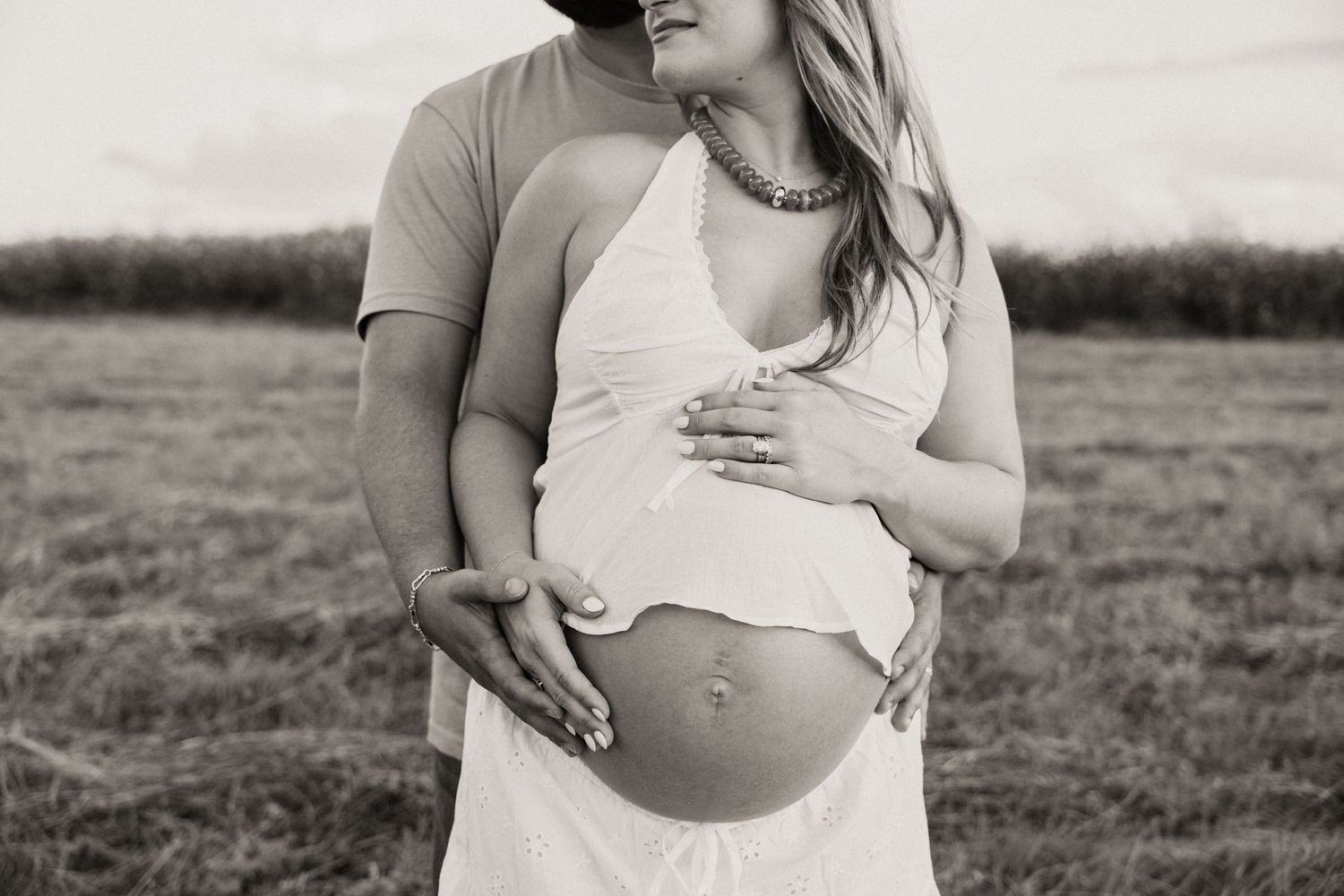 Black and white photo of a couple embracing outdoors, showing pregnant belly with hands tenderly placed during outdoor maternity session.