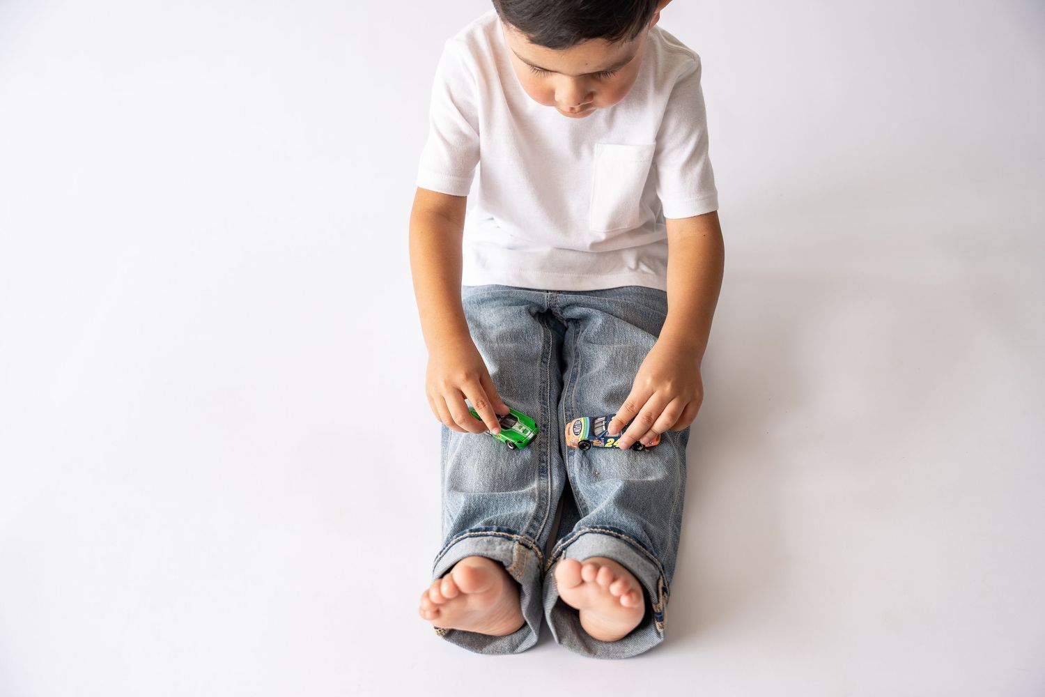 Young boy sitting on white background playing with small green toy cars in his hands.