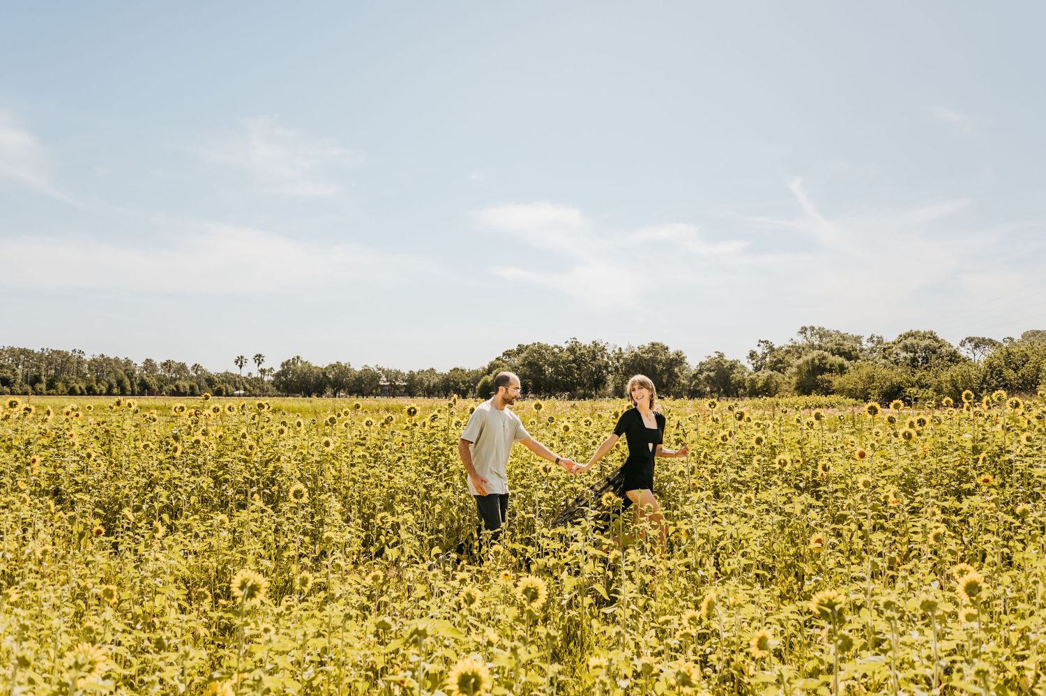 Two figures walk through a vibrant yellow field of wildflowers on a sunny day with trees in the background.