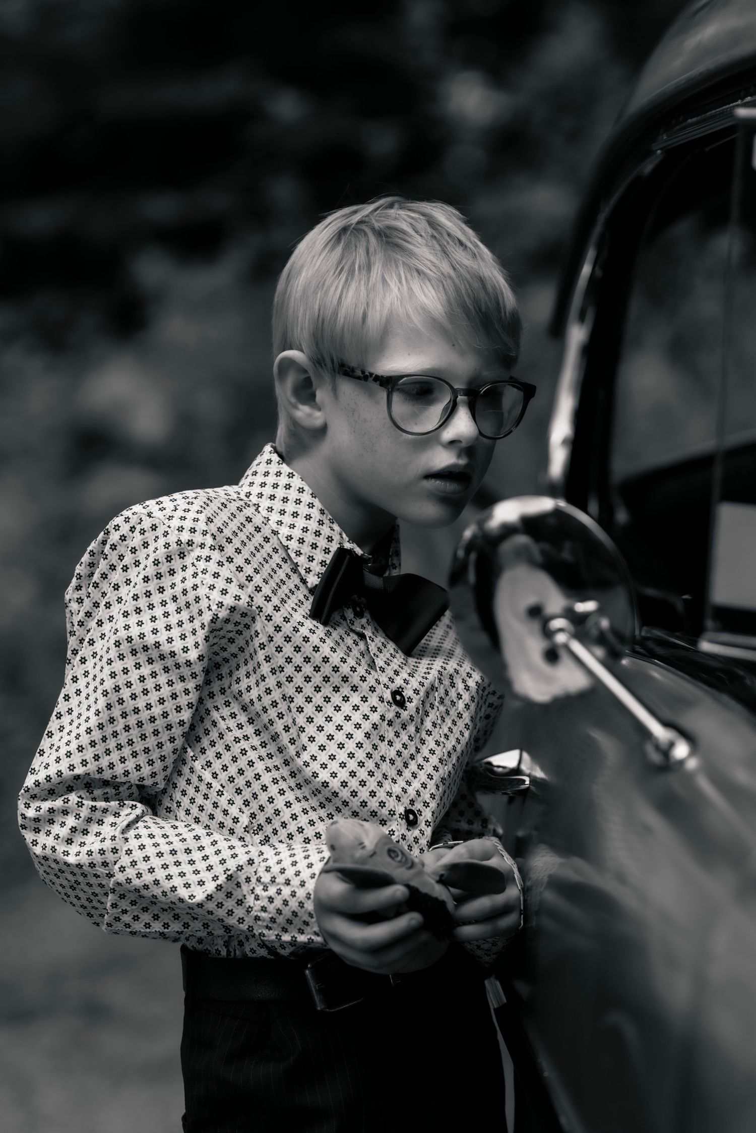 Retro styled black and white portrait of young boy in polka dot shirt and bowtie next to vintage car.