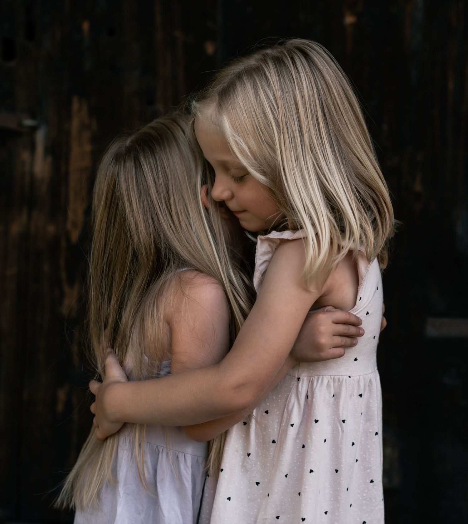 Intimate moment between two young girls in white dresses hugging against dark background.