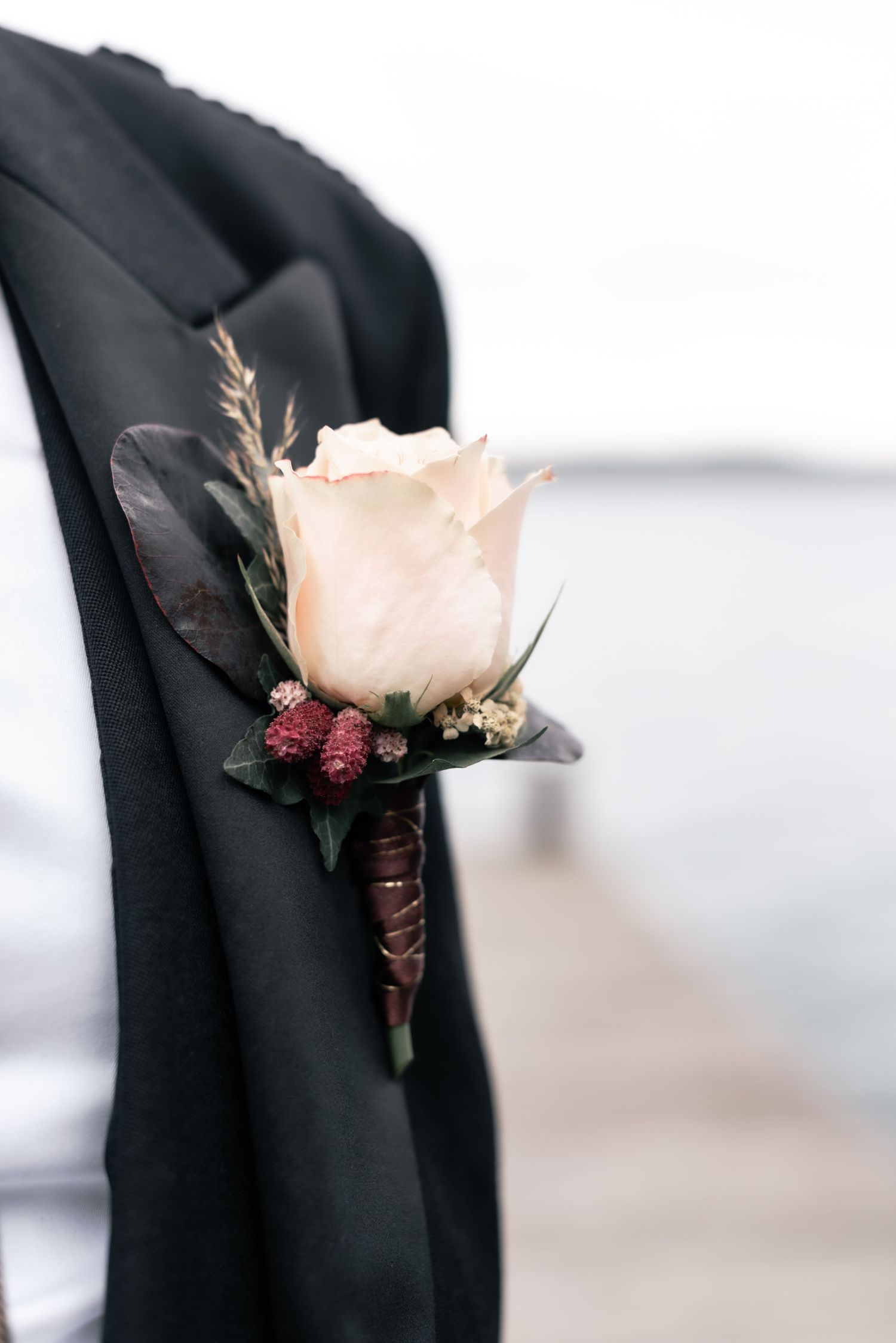 A delicate cream rose boutonniere with burgundy accents pinned to a black suit lapel.