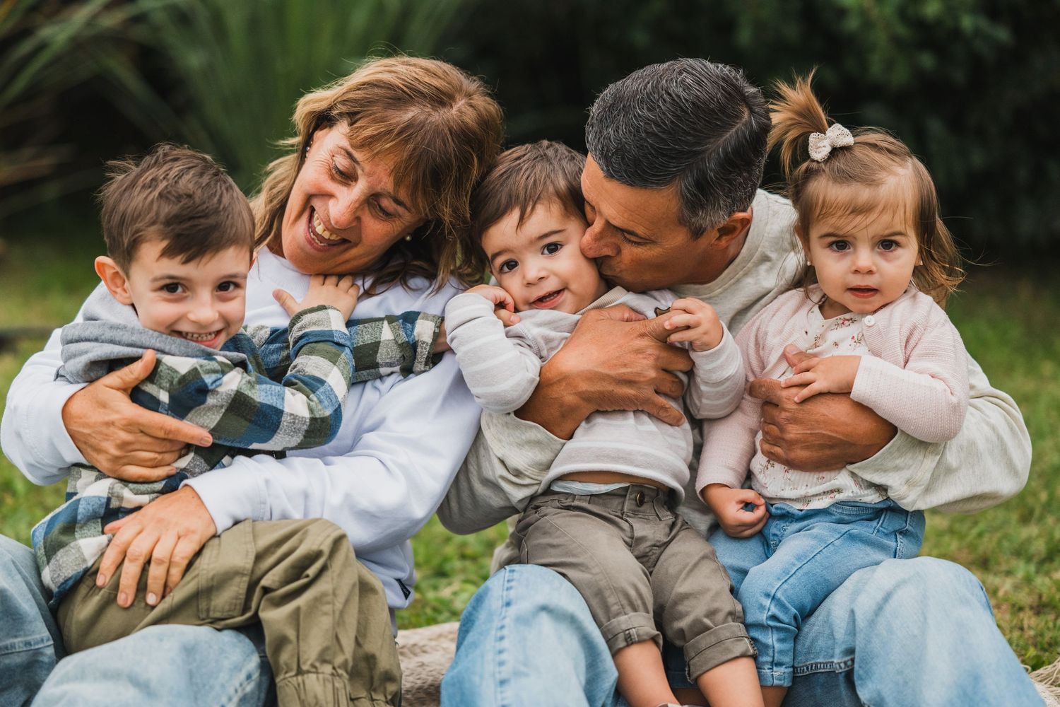 Abuelos sentados sobre lona en el pasto con sus tres nietos sobre su regazo, sonriendo y besándolos