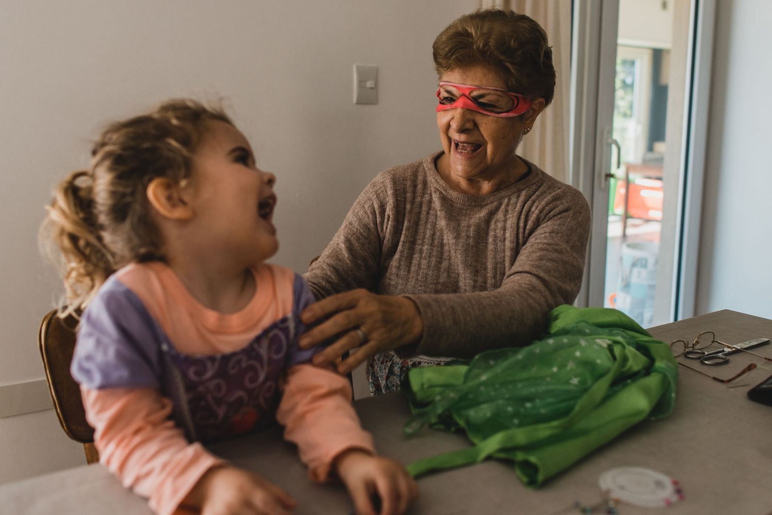 Abuela cosiendo disfraz de su nieta con antifaz puesto, la nieta la mira y se ríe