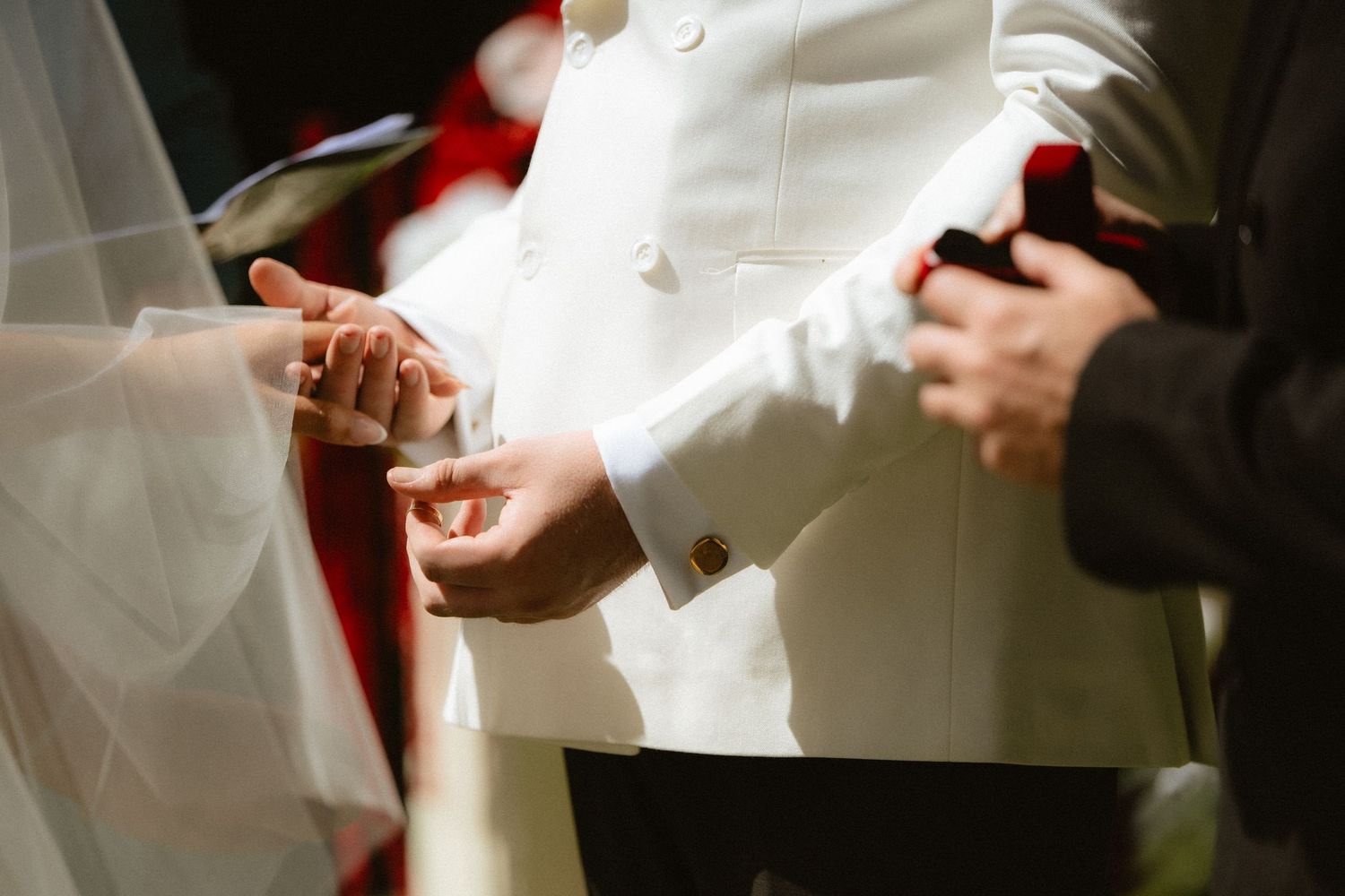 Groom in white suit placing wedding ring on bride's finger during ceremony.