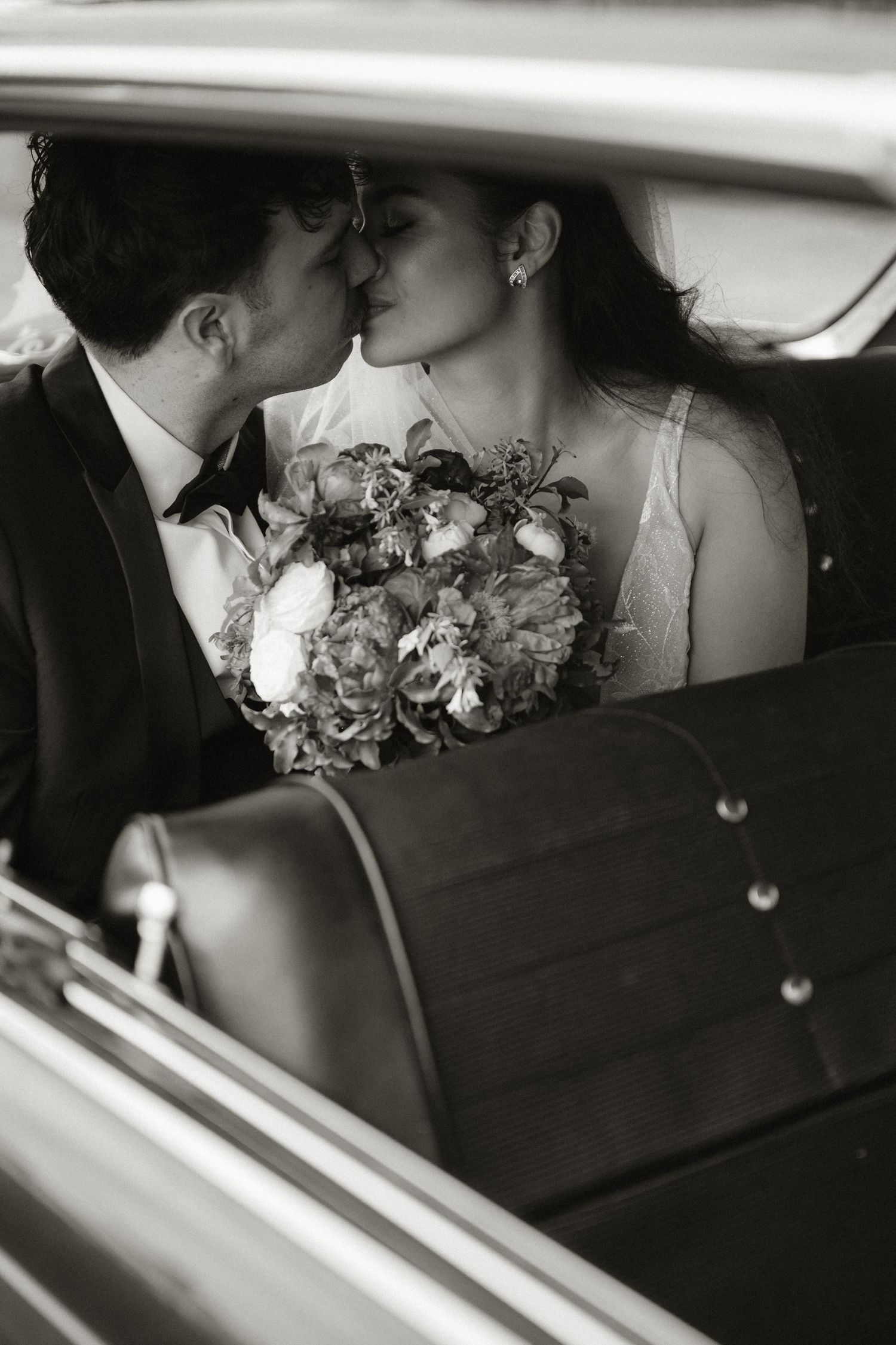 Black and white photo of a bride and groom sharing a kiss in the backseat of a vintage car, bouquet in hand.