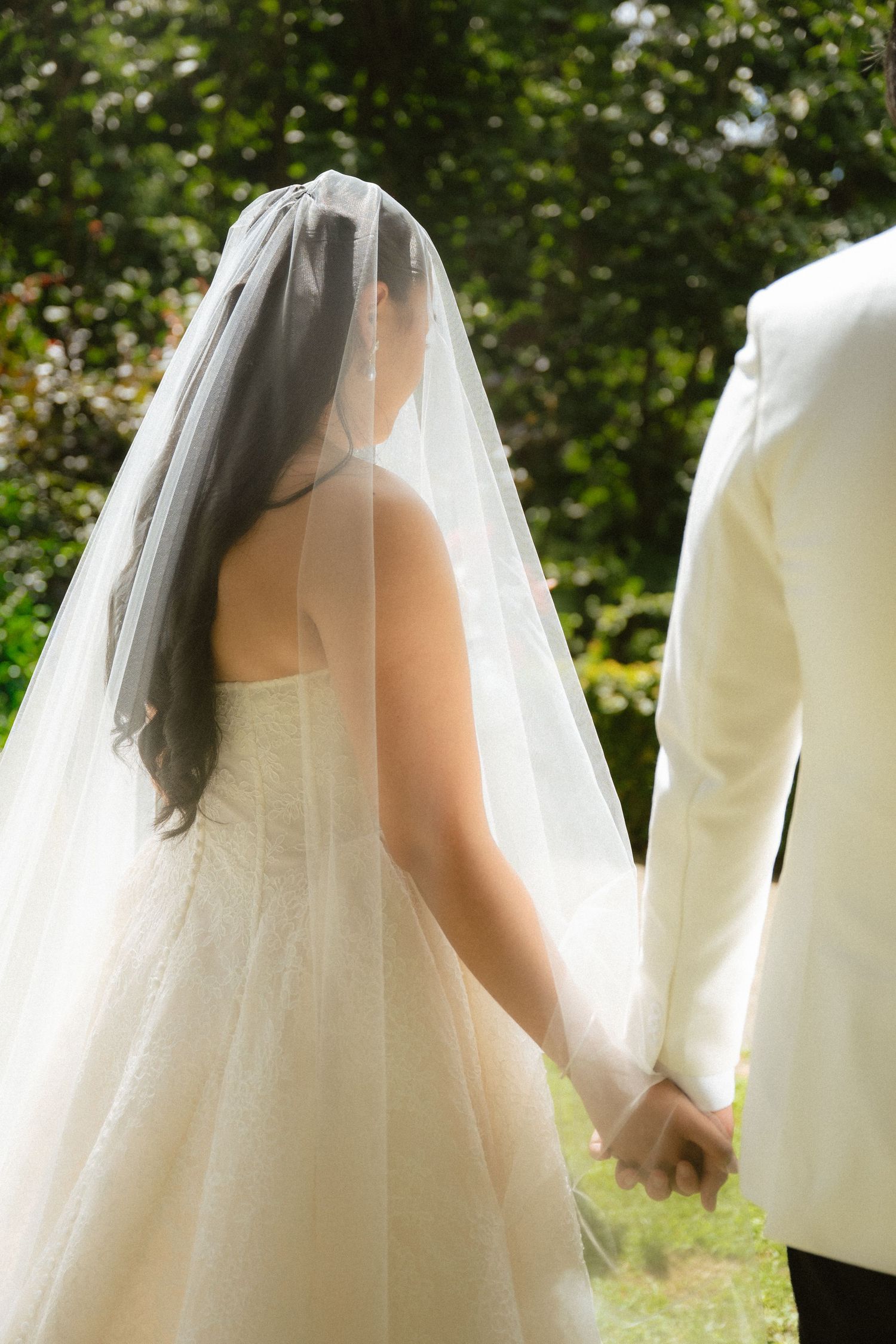 Bride in white gown and veil holds hands with groom in white suit, walking through lush green garden.